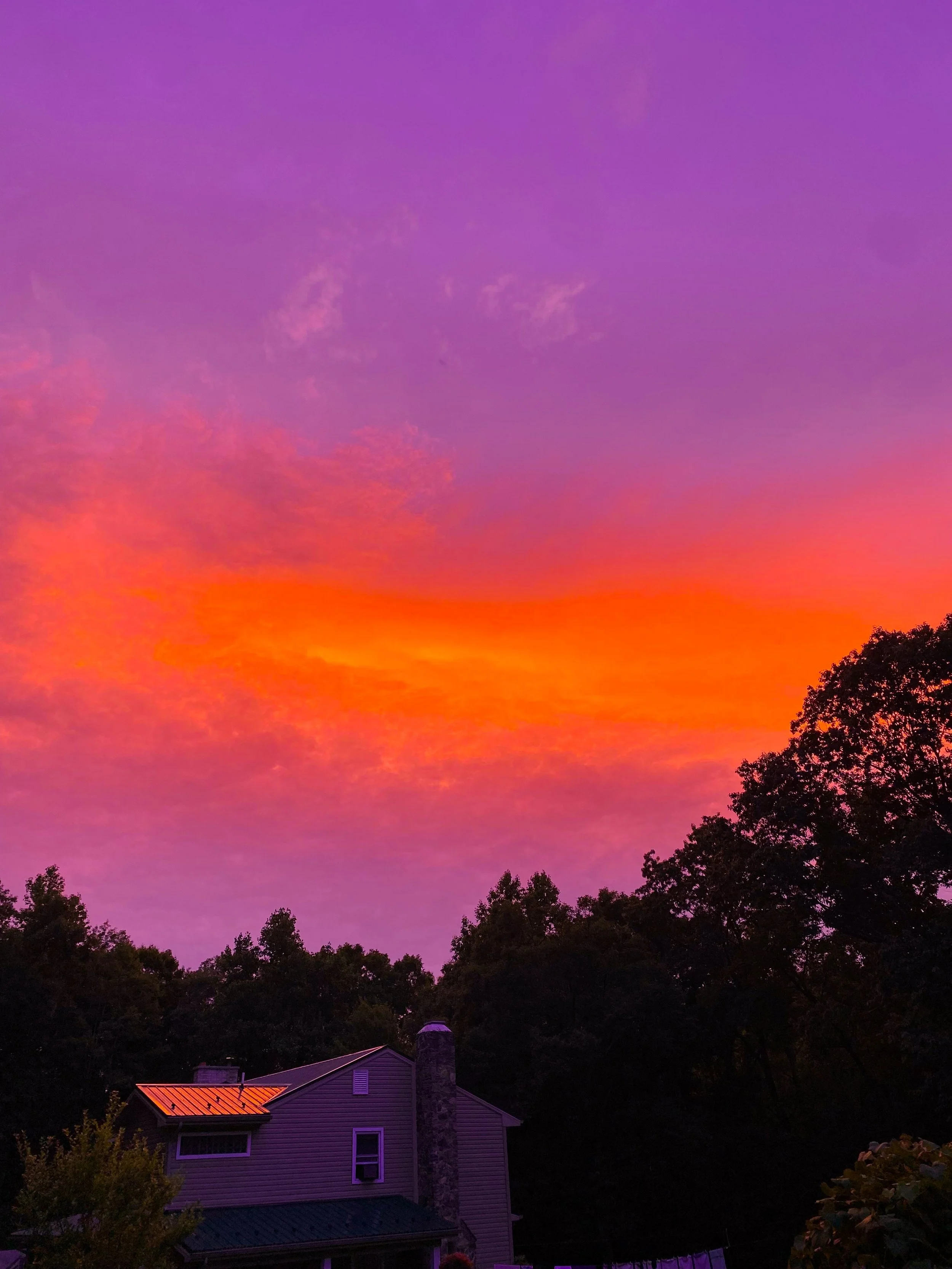 A colorful sunset sky with shades of purple, pink, orange, and yellow above a silhouette of trees and a house with a stone chimney.