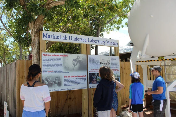 Four children observe an informational sign about MarineLab Undersea Laboratory Museum outdoors, with a wooden fence and trees in the background.