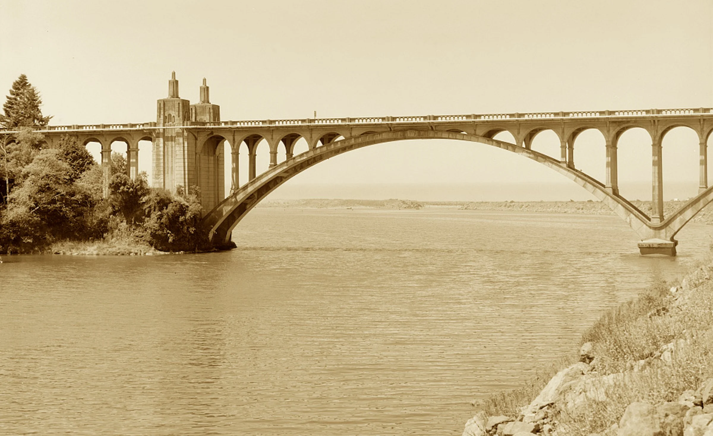 With arches that ripple in ribbed concrete span,
It carried the coast where no ferry boat ran.
McCullough designed with an Art Deco flare,
A bridge light and slender that hangs in sea air.
The first of its kind, where the Rogue meets the tide—
An ele