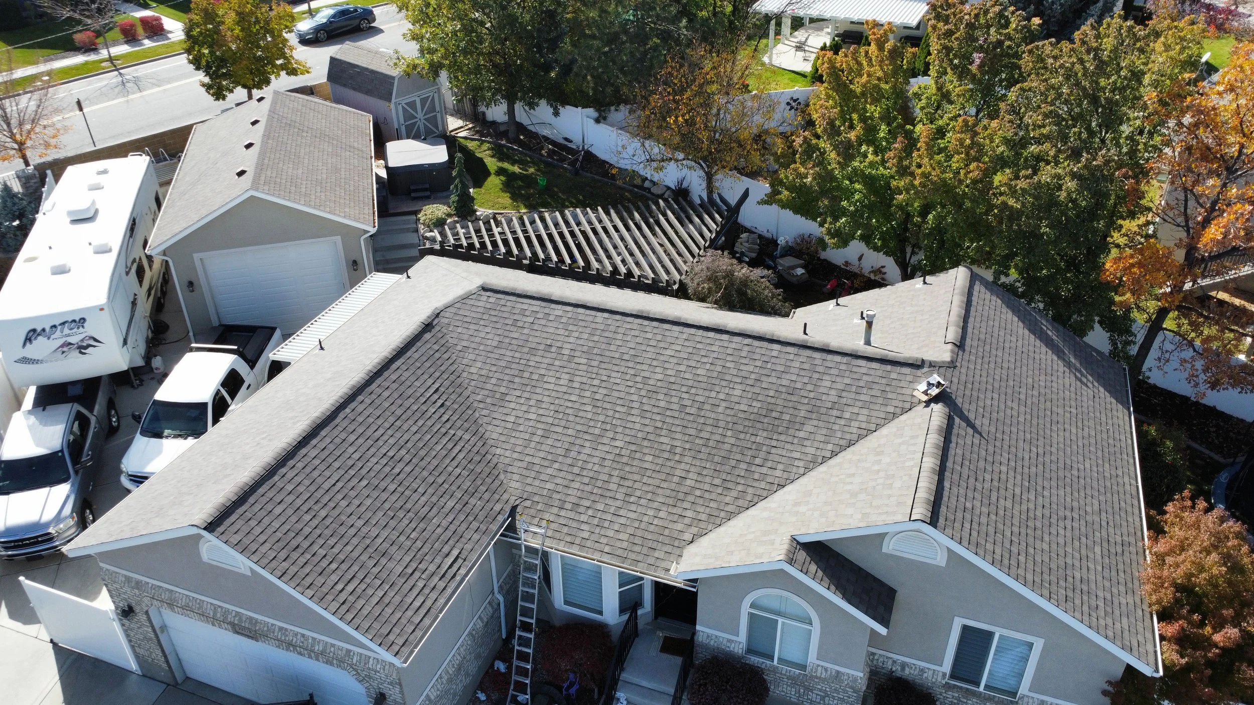 An aerial view of a residential backyard showing a gray-shingled roof, a white house, a landscaped area with trees and plants, a deck area, and several parked cars in the driveway.