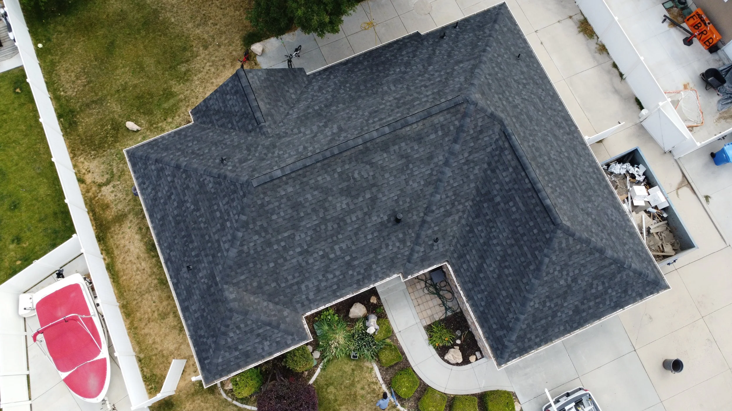 An aerial view of a house with a dark shingle roof, a backyard with a green lawn, patio, a trampoline, and various items including a basketball hoop, garden beds, and construction debris.