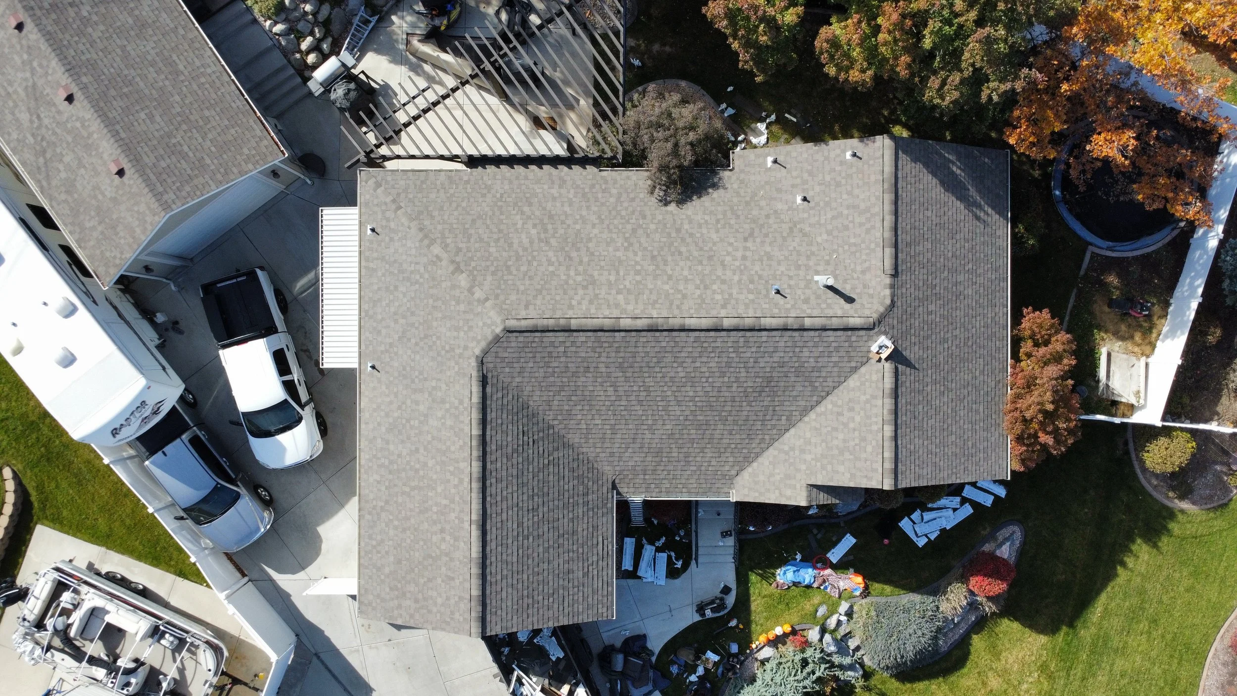 An aerial view of a house with a grey shingle roof and a backyard that has trees with fall-colored leaves, a trampoline, and circular pathways. There are multiple parked vehicles in the driveway, including trucks and SUVs, and some equipment and furn