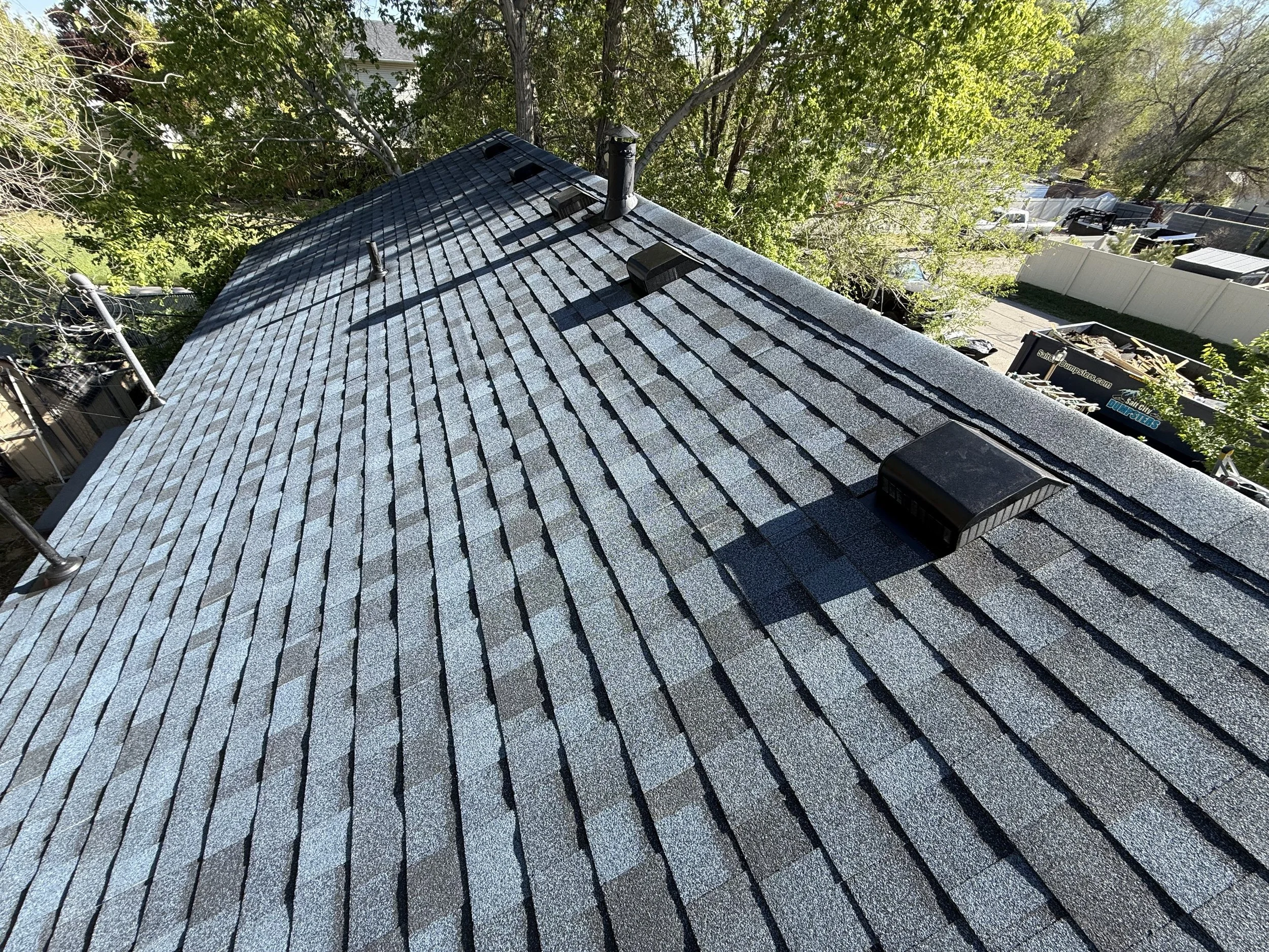 View of a rooftops covered with gray asphalt shingles, trees with green leaves in the background, a white fence, and a truck parked on a driveway.