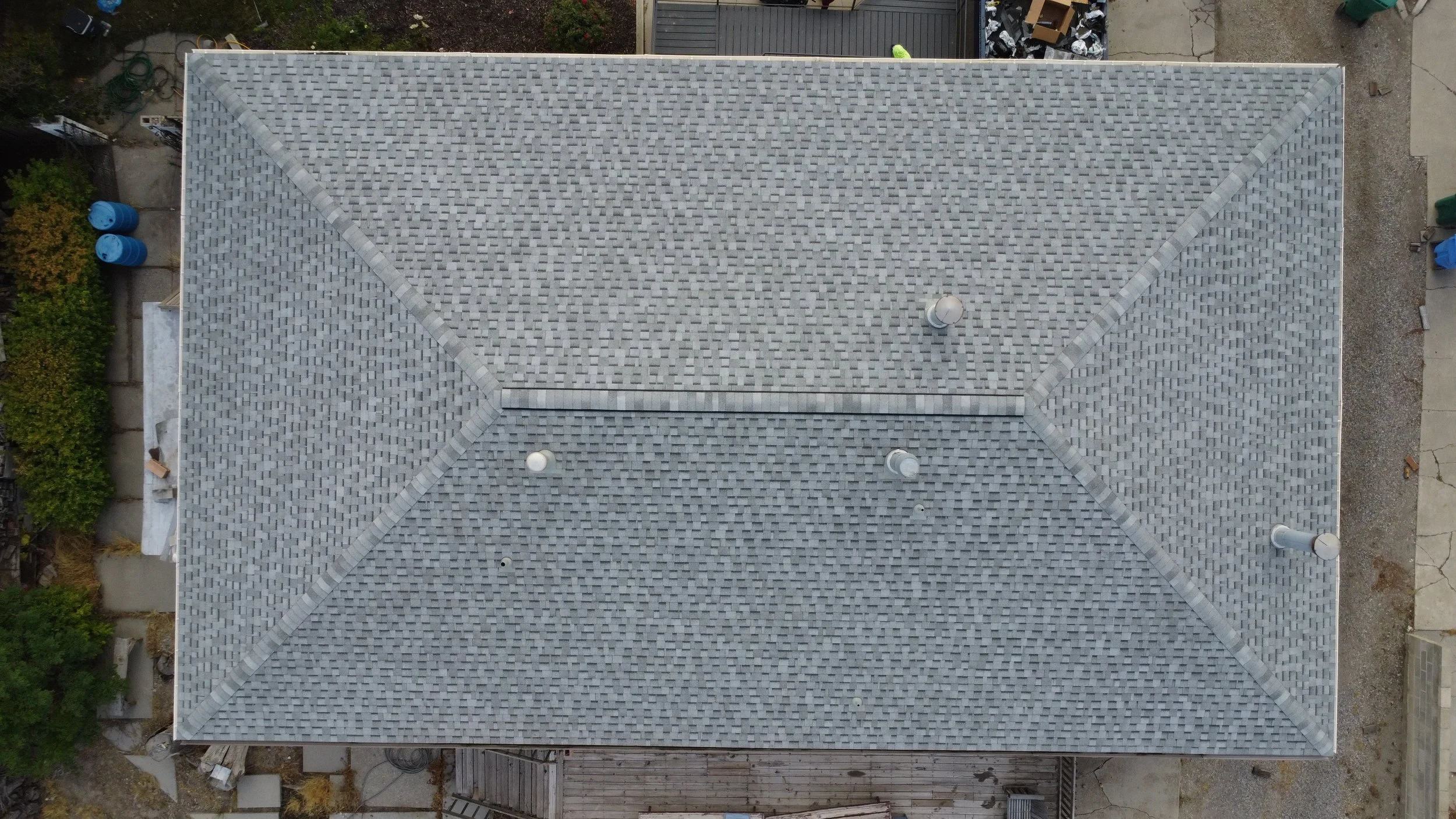An aerial view of a house roof with gray shingles and several vents, surrounded by a backyard with trees, a fence, and miscellaneous backyard objects.