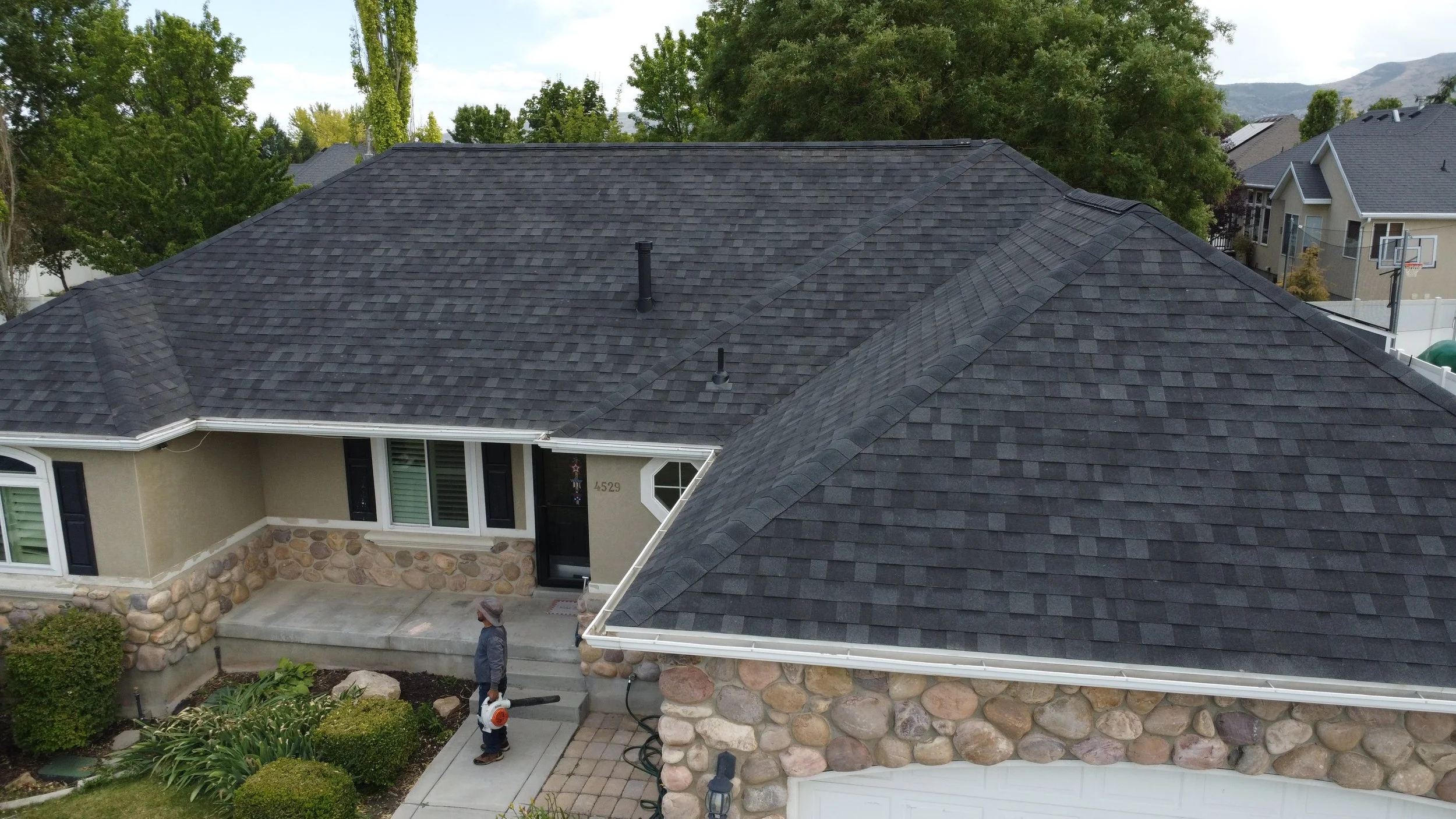 Aerial view of a house with a dark gray shingle roof, trees, neighboring houses, and a mountain backdrop. Two people are working on the front yard, one with a leaf blower.
