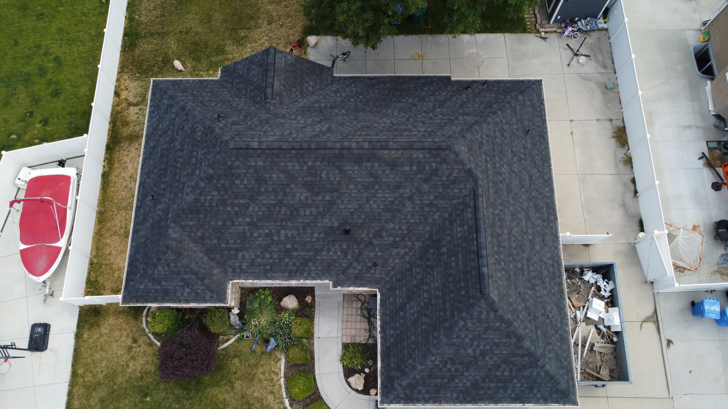 An aerial view of a house with a dark shingled roof, a backyard with a concrete patio, bicycles, and various items, and a boat on a trailer.