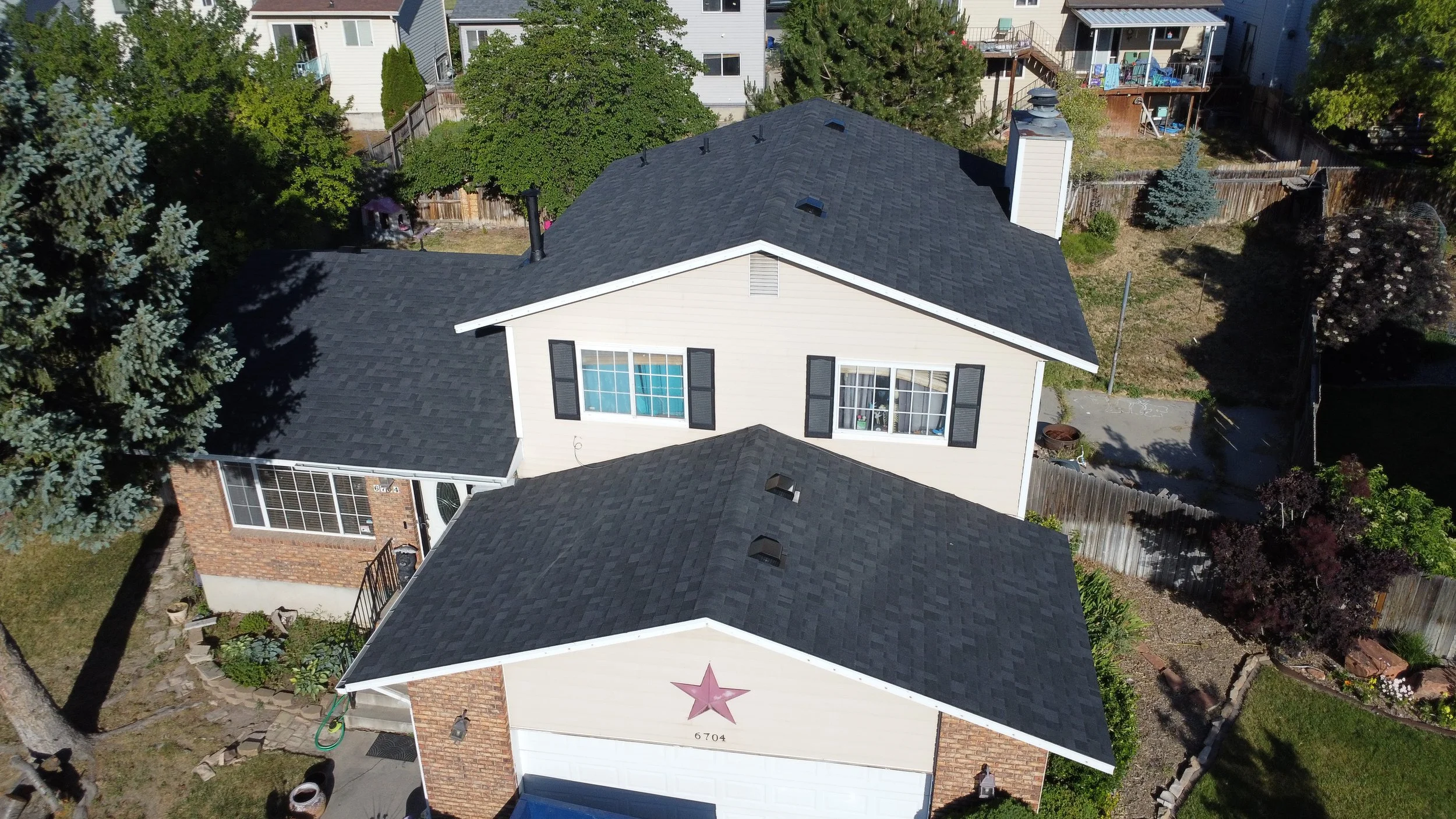 Aerial view of a suburban house with a black shingle roof, beige siding, black shutters, and large windows in a yard with trees, plants, and neighboring houses.