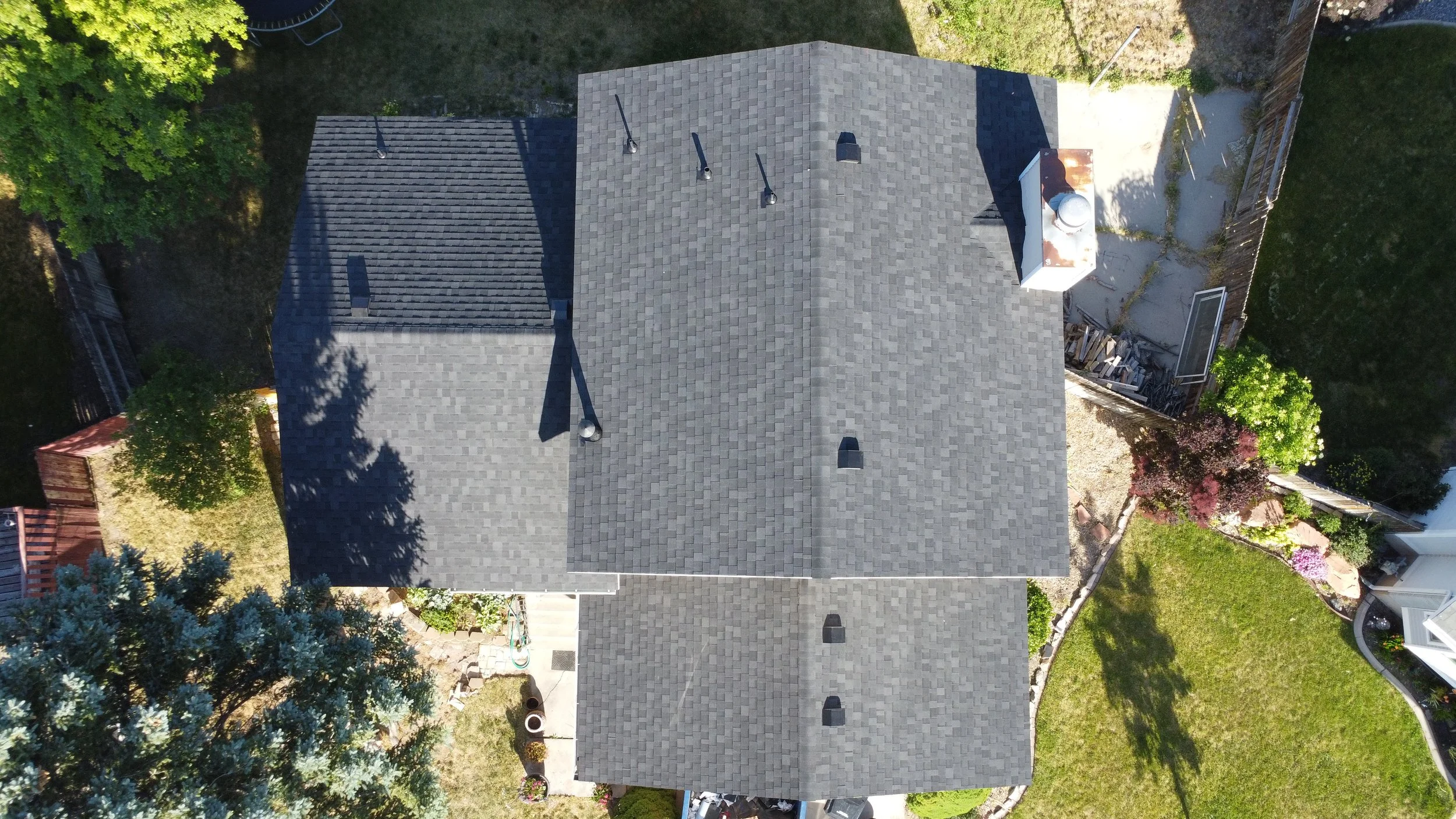 Aerial view of a gray-shingled house with landscaped yard, trees, and neighboring houses visible surrounding it.