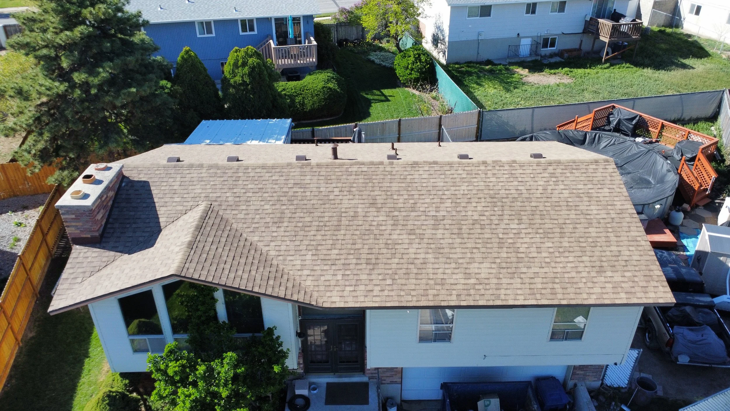 Aerial view of a suburban backyard with a house, a chimney, trees, a grassy area, a wooden deck, a fence, and a covered hot tub surrounded by a black tarp.