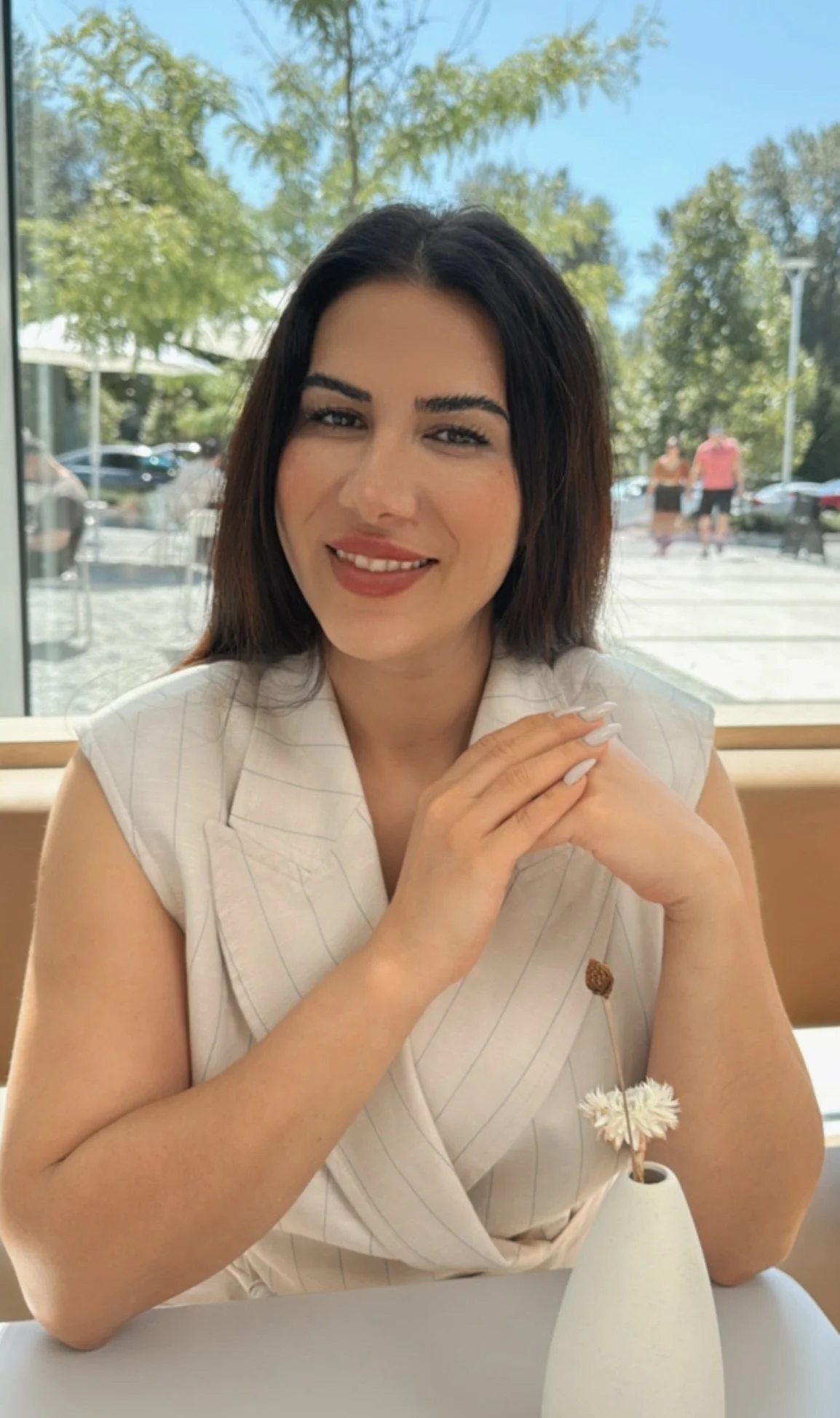 A smiling woman with dark hair and light makeup sitting at a table in a restaurant, with a view of trees and parked cars outside through large windows, holding her hands together near her face.