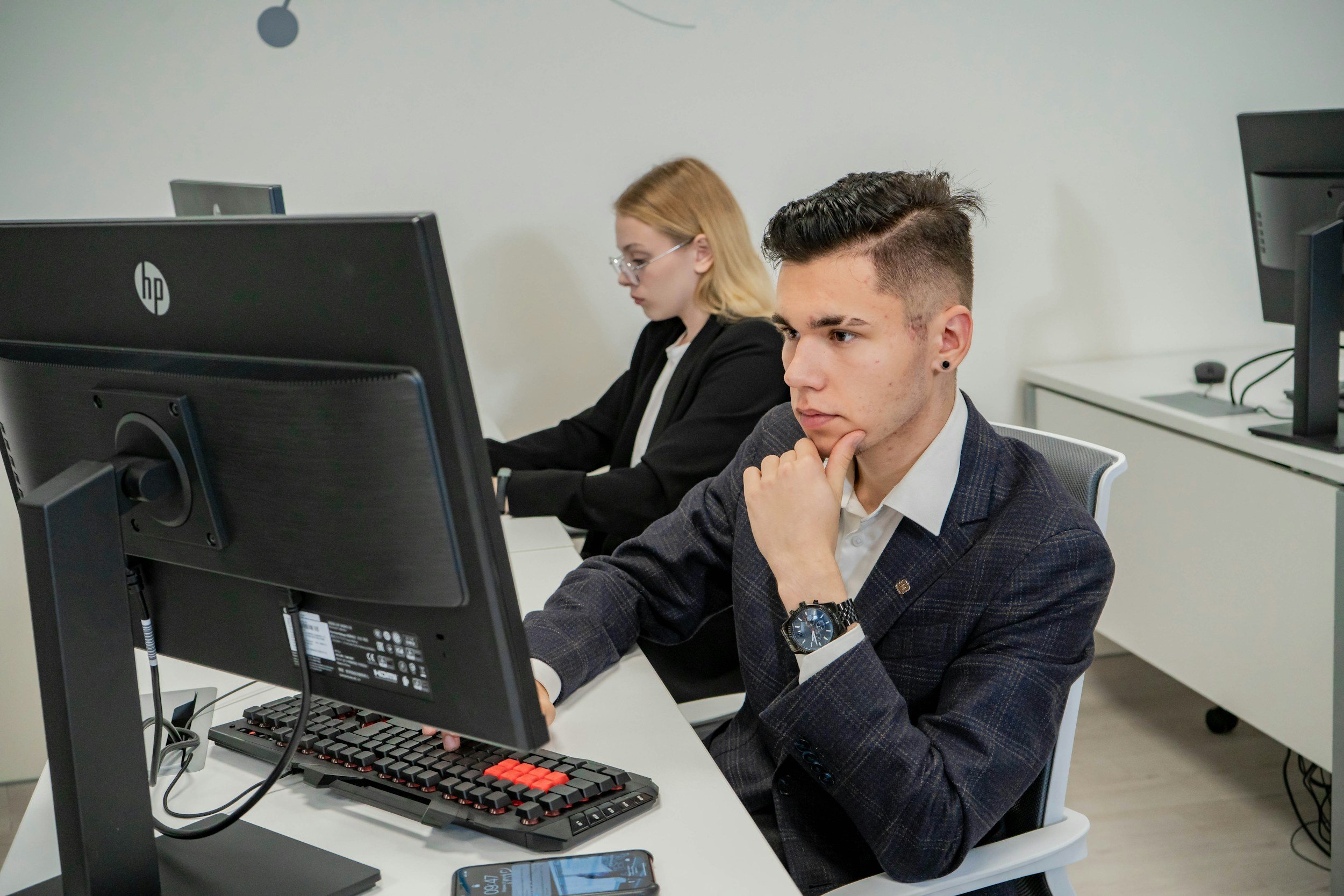 A man in a business suit sitting at a desk, looking at a computer monitor with a focused expression. A woman in a business suit is working at her desk in the background.