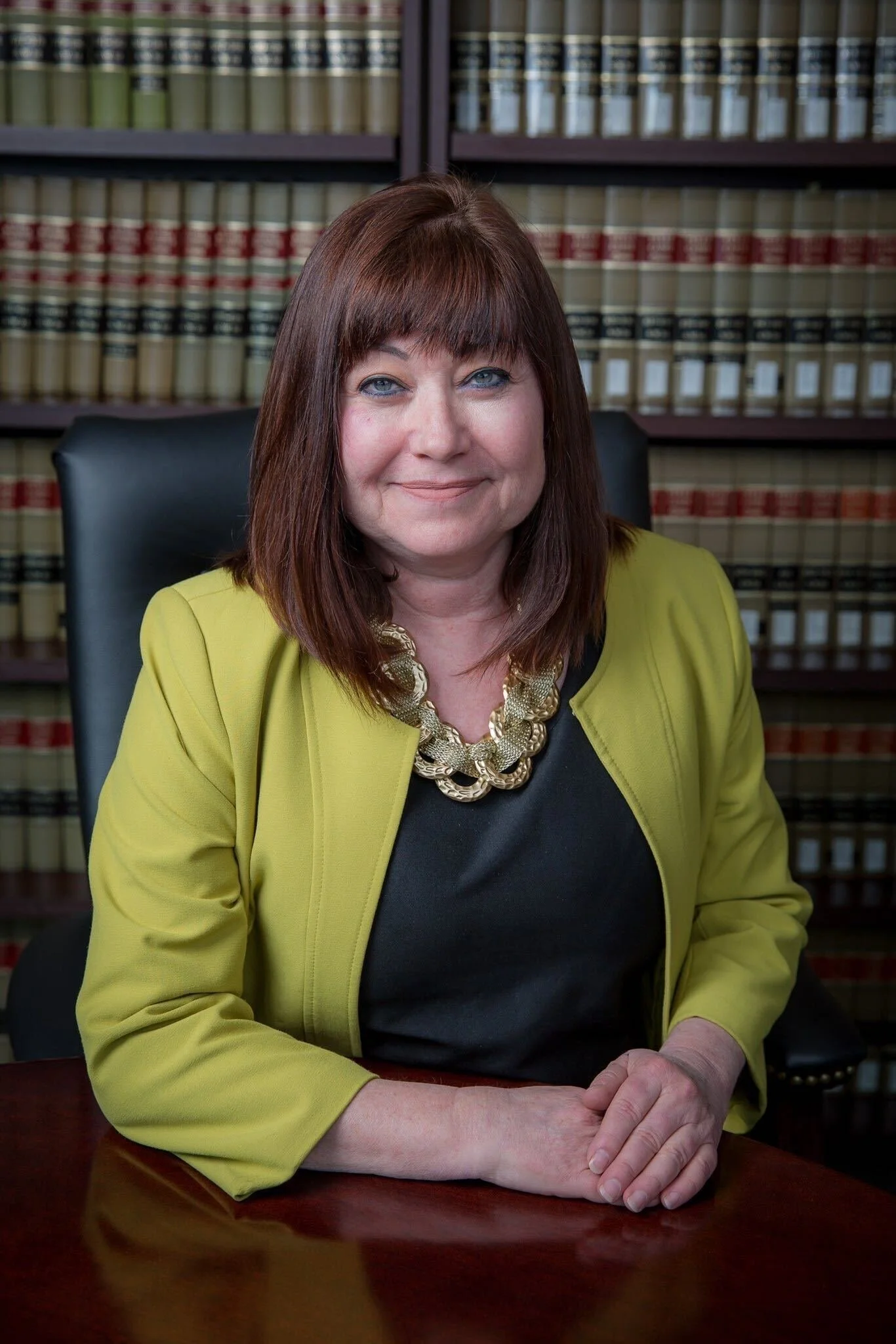 A woman with shoulder-length brown hair and blue eyes, wearing a yellow blazer, black top, and large gold chain necklace, sitting at a wooden desk with a bookshelf filled with law books in the background.