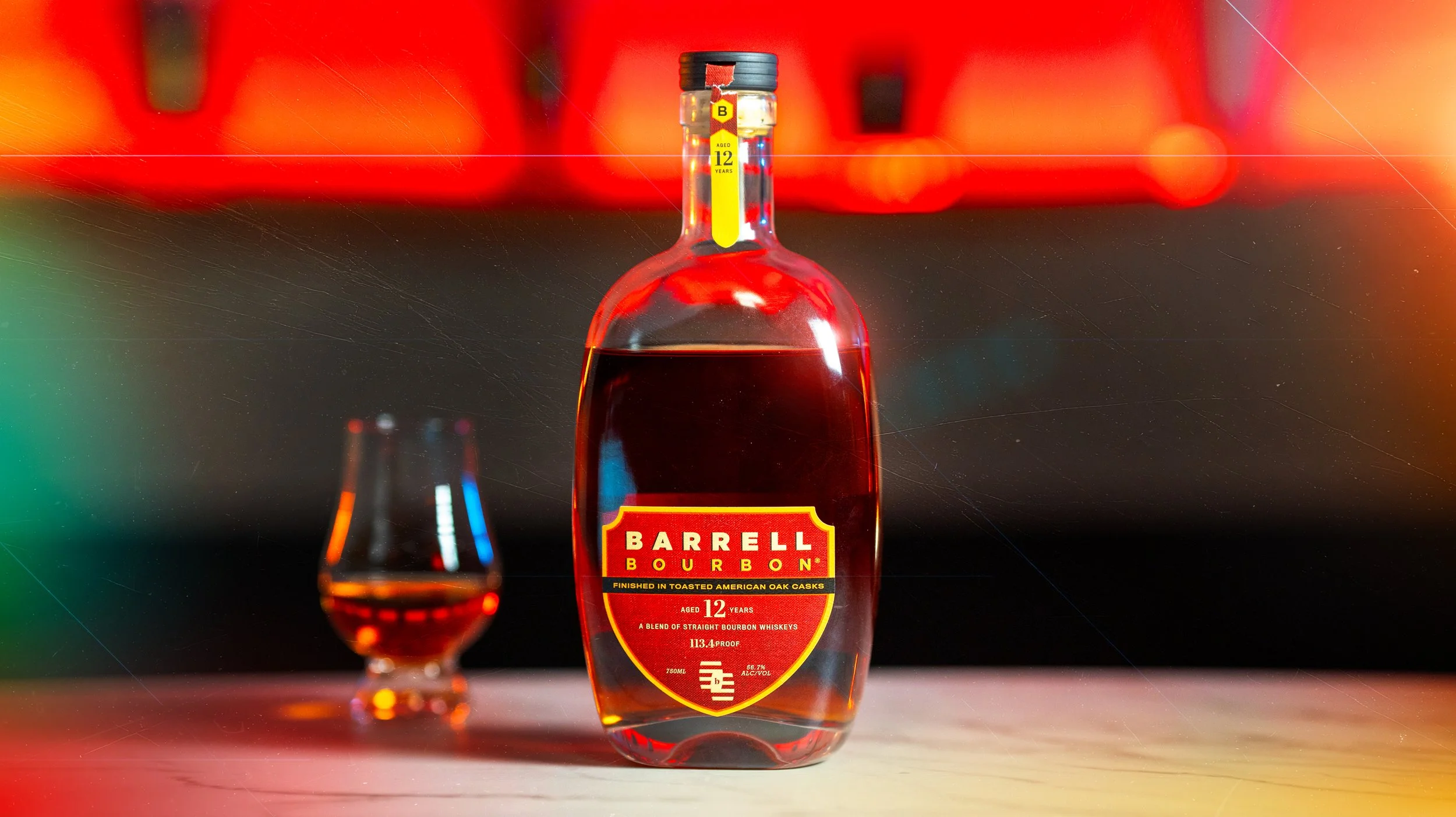 A bottle of Barrell Bourbon whiskey with red liquid inside, and a glass filled with the same red liquid in front of it, on a flat surface with blurry colorful background.