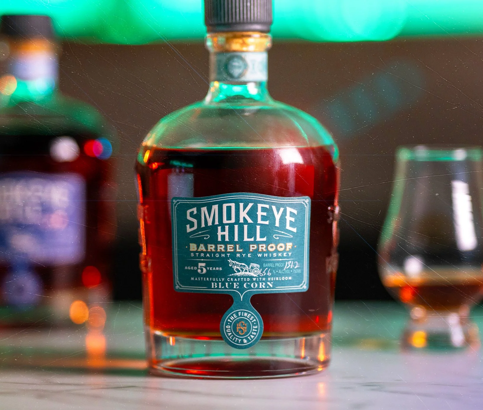 A bottle of Smokey Hill Barrel Proof straight rye whiskey with a blue label, placed on a bar counter with a glass of whiskey in the background.