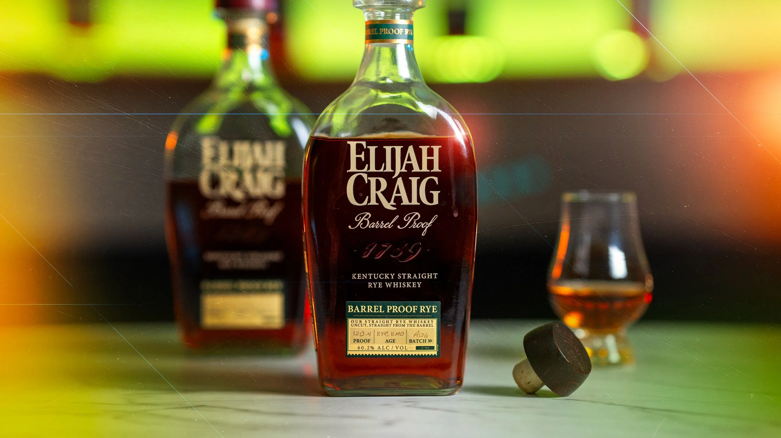 A bottle of Elijah Craig Barrel Proof Rye whiskey on a surface, with a glass and other whiskey bottles blurred in the background.