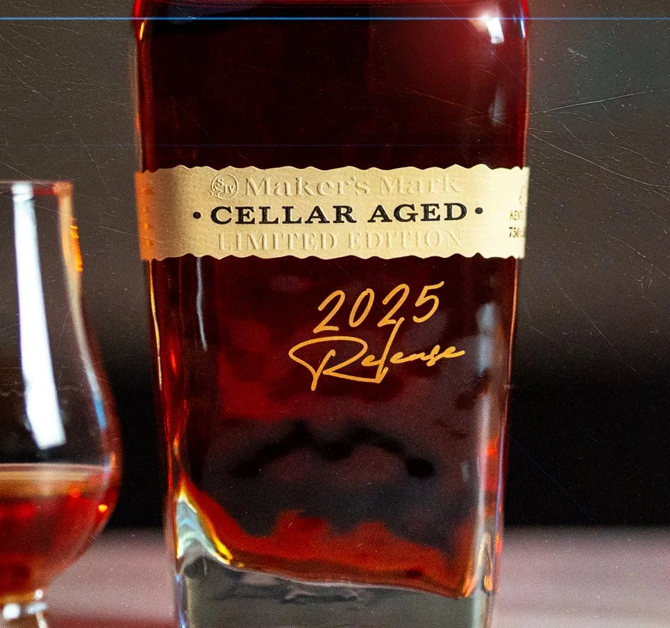 A bottle of Maker's Mark limited edition 2025 cellar aged whiskey with a gold label and signature, placed on a dark surface.