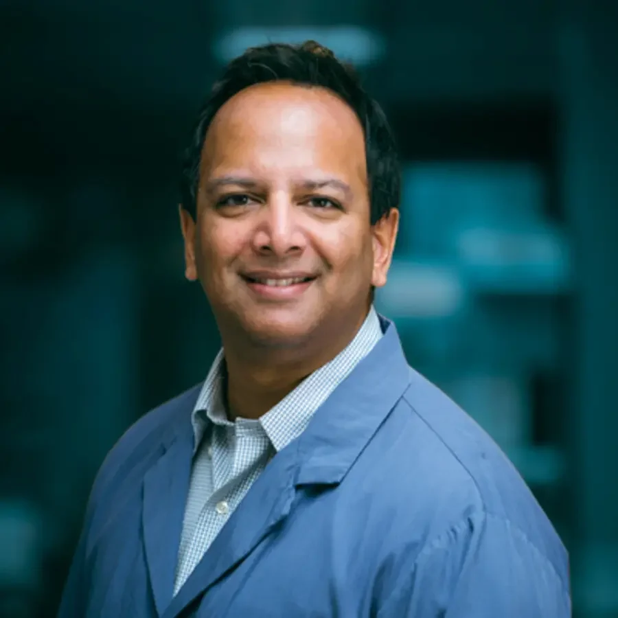 A smiling man in a light blue blazer and white shirt, standing outdoors in an urban setting.