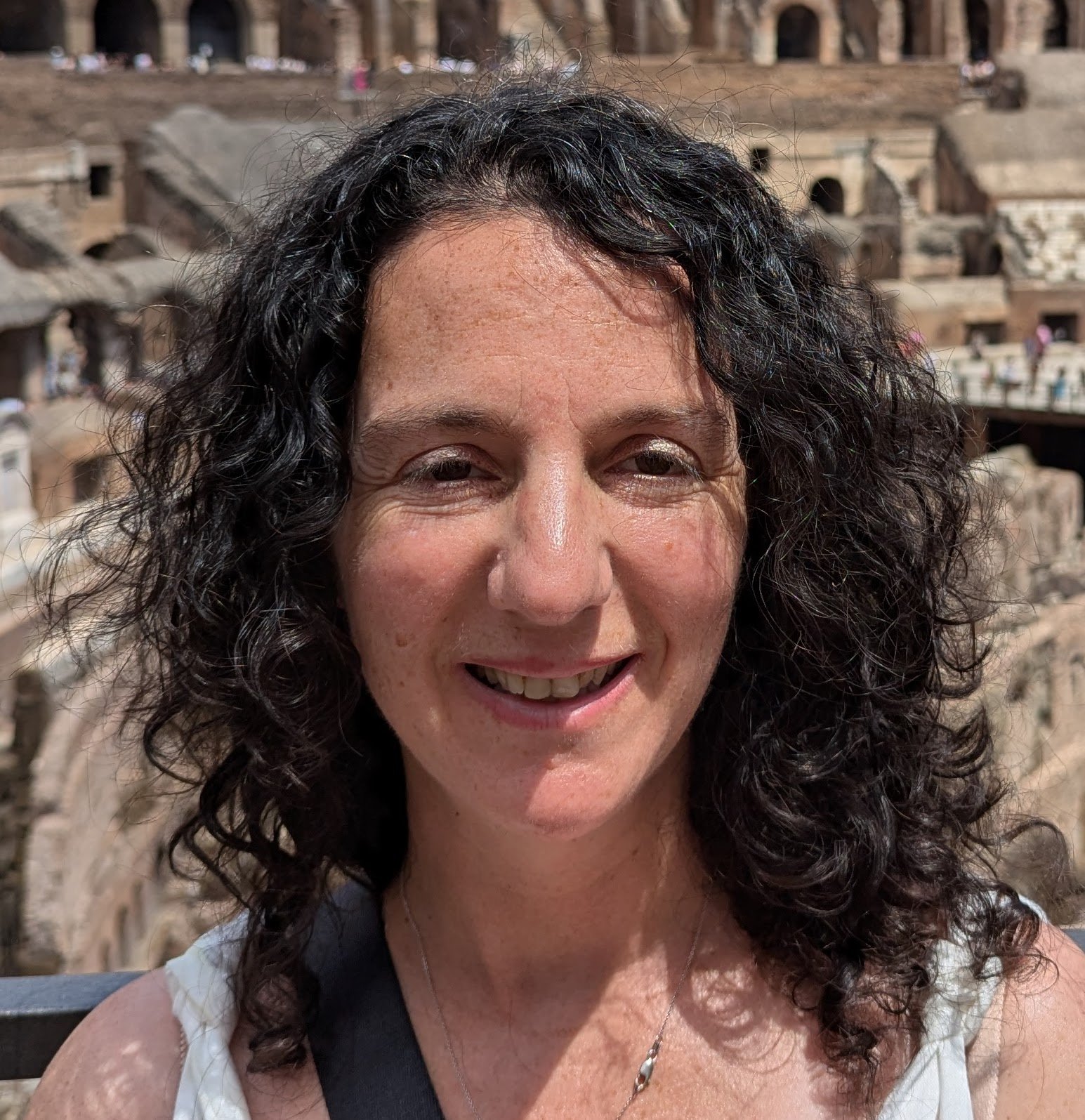 A woman with curly dark hair smiling outdoors with ancient ruins in the background.