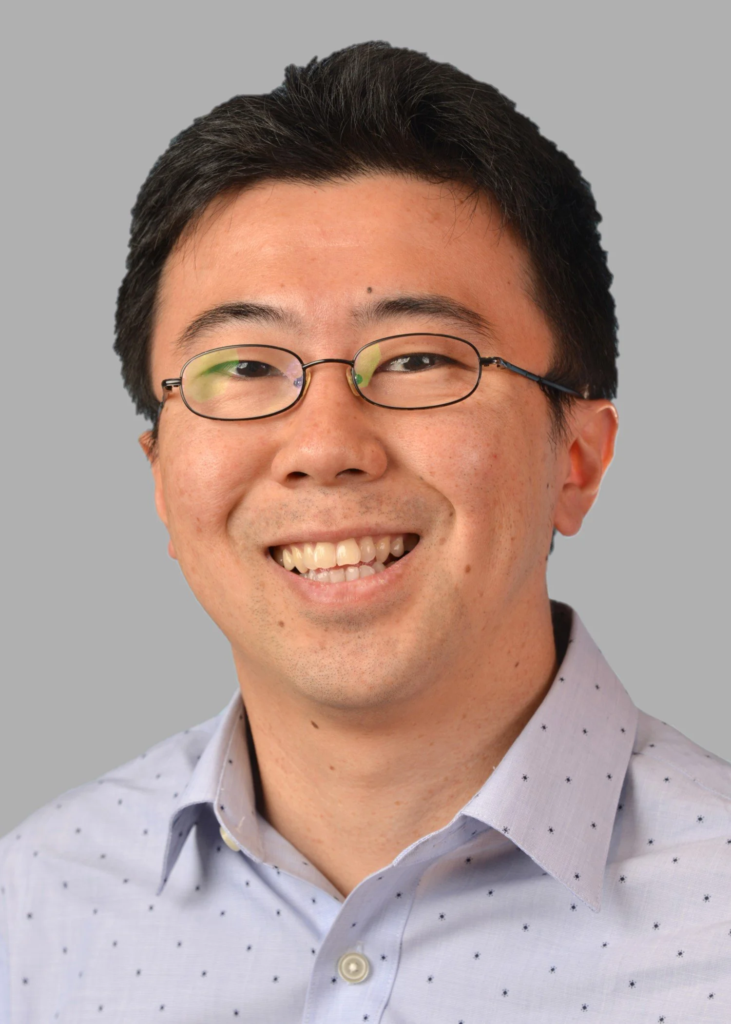 Portrait of a smiling man with glasses and short dark hair, wearing a light blue dress shirt with a small dot pattern, against a plain gray background.