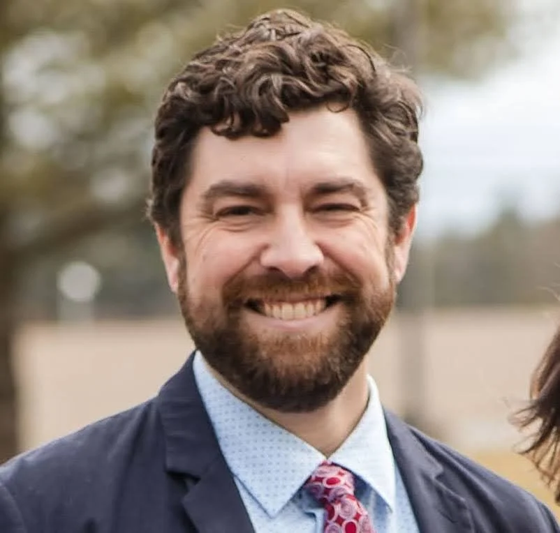 A man with curly dark hair, glasses, a beard, and a nose ring wearing a dark green sweater, sitting indoors.