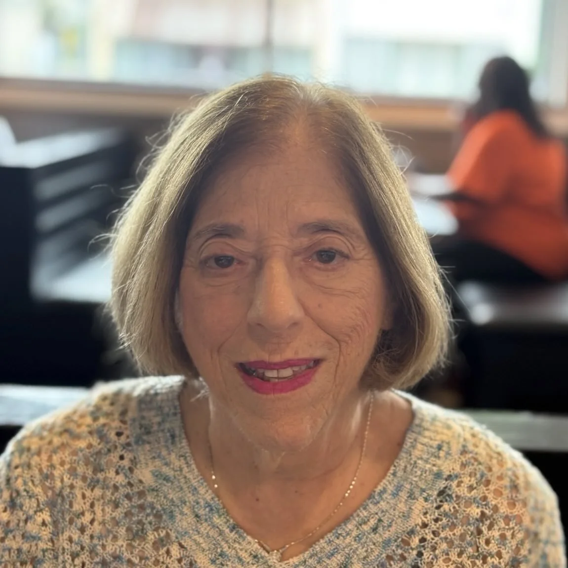 A smiling woman with shoulder-length black hair wearing a blue top outdoors with green foliage in the background.