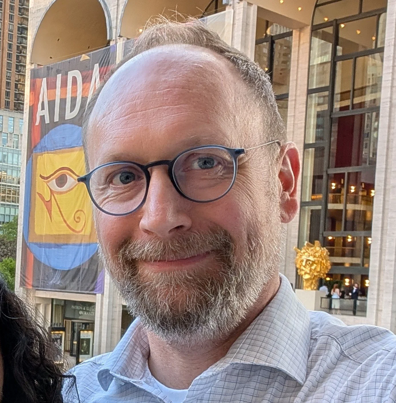 Close-up of a smiling man with glasses and a beard, standing outside in front of a building with a large colorful banner and a golden art piece, in an urban setting.
