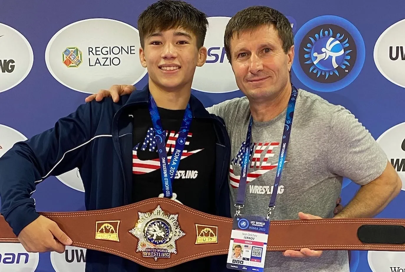 Two men smiling, one young and one older, holding a large wrestling championship belt in front of a backdrop with logos, including 'Regione Lazio' and 'UWW World Championship Rome 2022'.