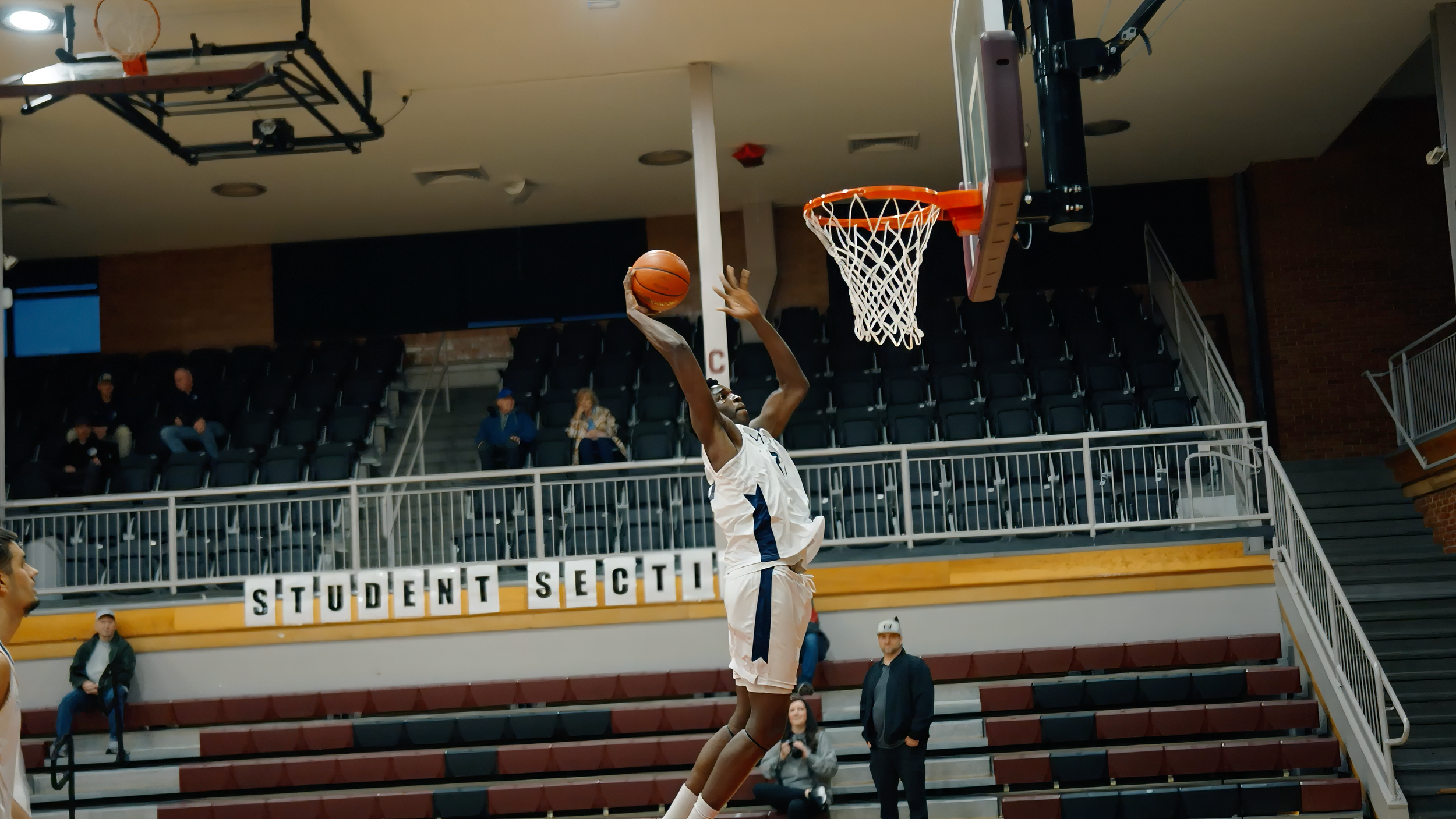 A basketball player jumping to make a shot in an indoor basketball court, with spectators seated in the background and a sign reading 'STUDENT SECTI' on the wall.