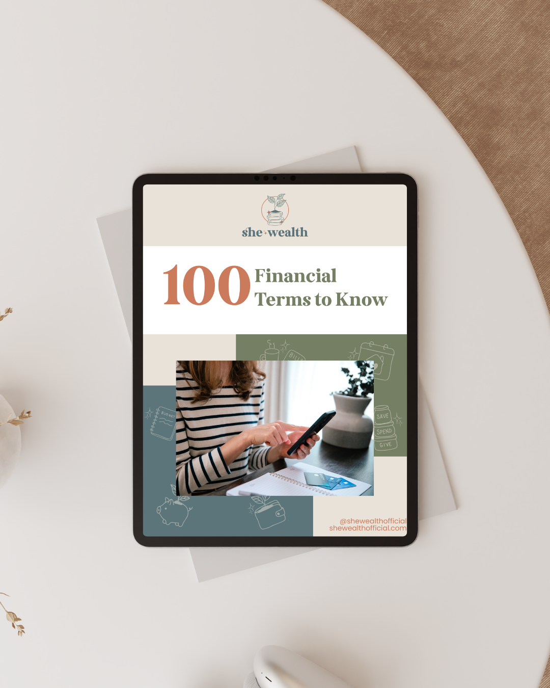 An iPad on a white table displaying an e-book titled "100 Financial Terms to Know" by she•wealth. The cover features a woman in a striped shirt using a smartphone, with the company's logo and social media handle at the bottom right corner.