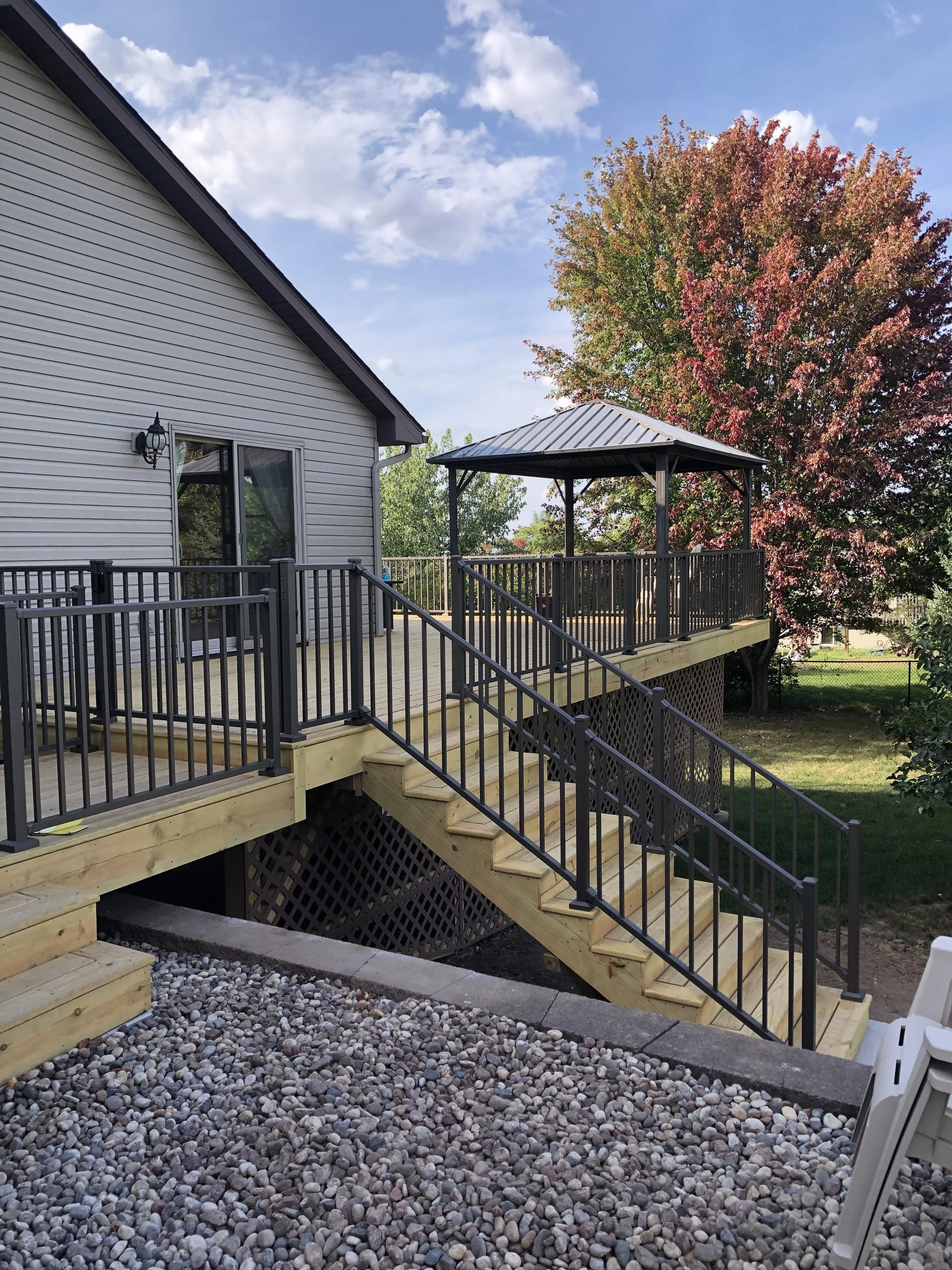 A newly built wooden deck with a staircase, black metal railing, and a covered gazebo area, attached to a house with white siding. The scene is outdoors with a large tree with red and green foliage, blue sky, and scattered clouds.