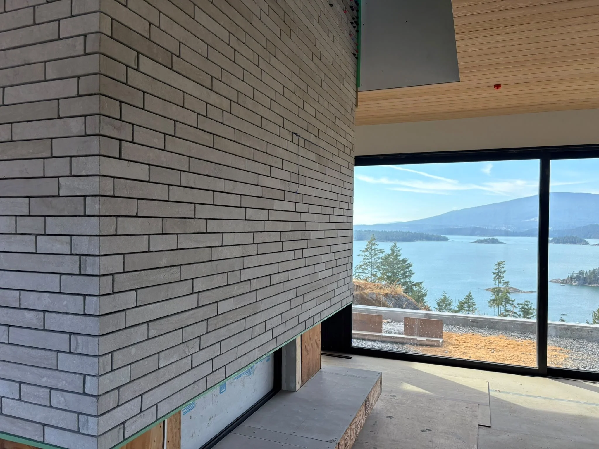 Interior of a modern house under construction with a brick wall, wooden ceiling, and large window overlooking a lake and mountains.