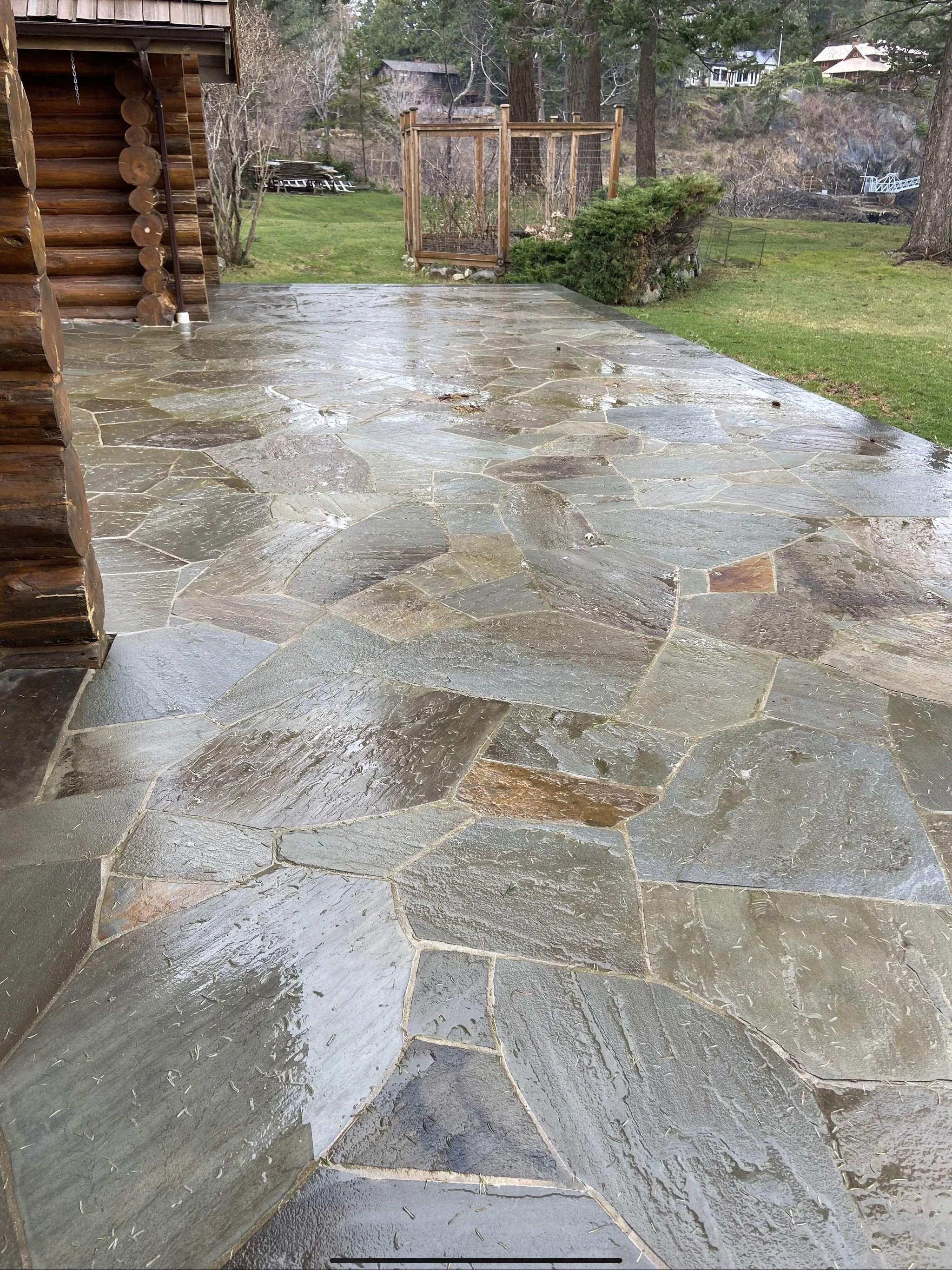 Wet, polished stone patio outside a wooden house with a lush green yard and trees in the background.