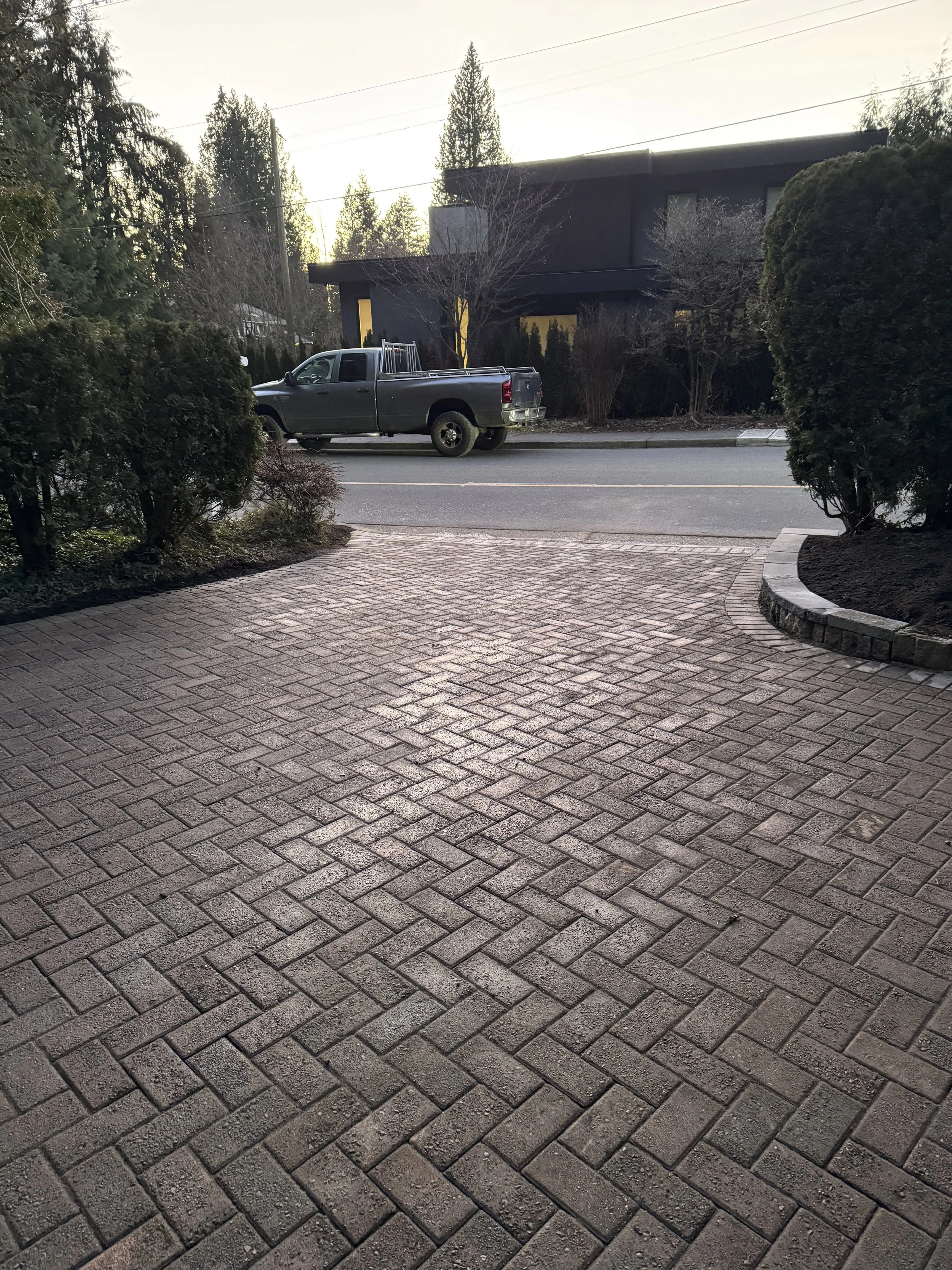 View of a paved brick driveway leading to a street with a dark gray pickup truck parked along the curb. There are bushes and trees on both sides of the driveway, and a modern black building in the background with leafless trees and power lines visibl