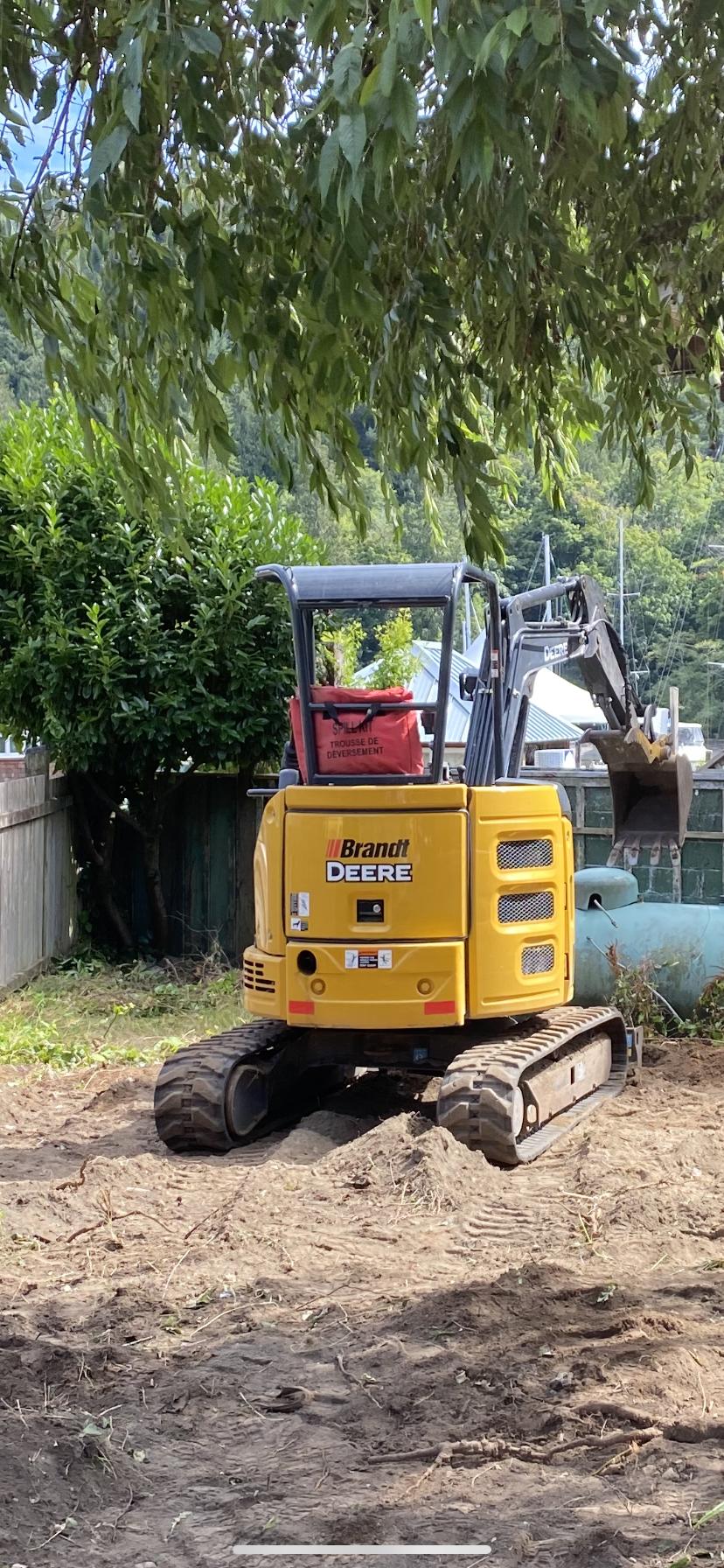 A yellow mini excavator with tracks, branded 'Brandt Deere,' is positioned on a dirt patch in a backyard, with a large tree overhead and a fence and propane tank in the background.