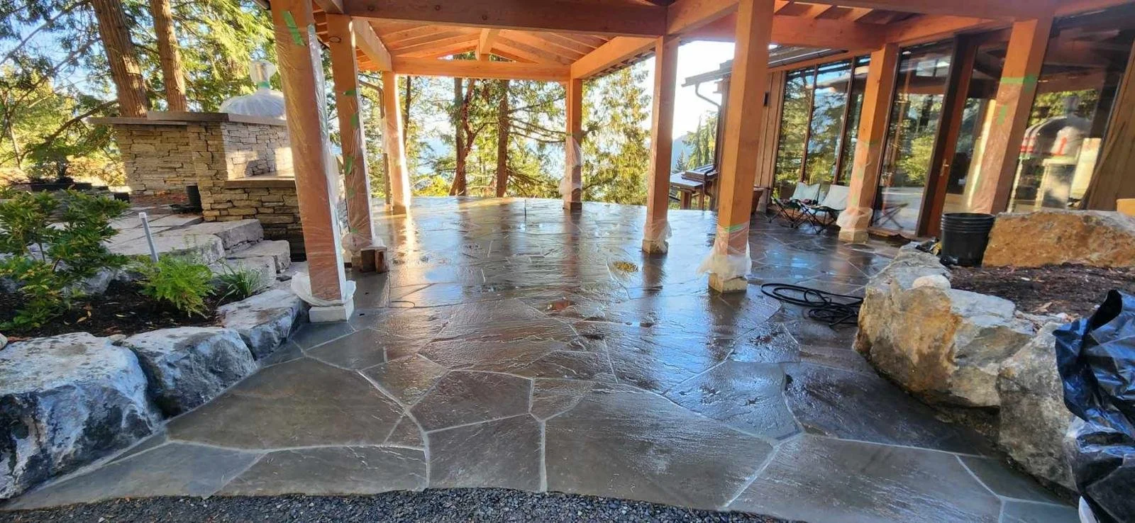 Covered outdoor patio with a stone and concrete floor, surrounded by trees, with a built-in stone oven or fireplace on the left and some outdoor furniture on the right, still under construction.