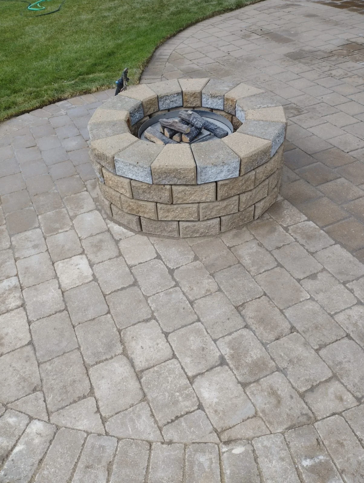 A fire pit surrounded by bricks on a stone patio, with a grassy area nearby.