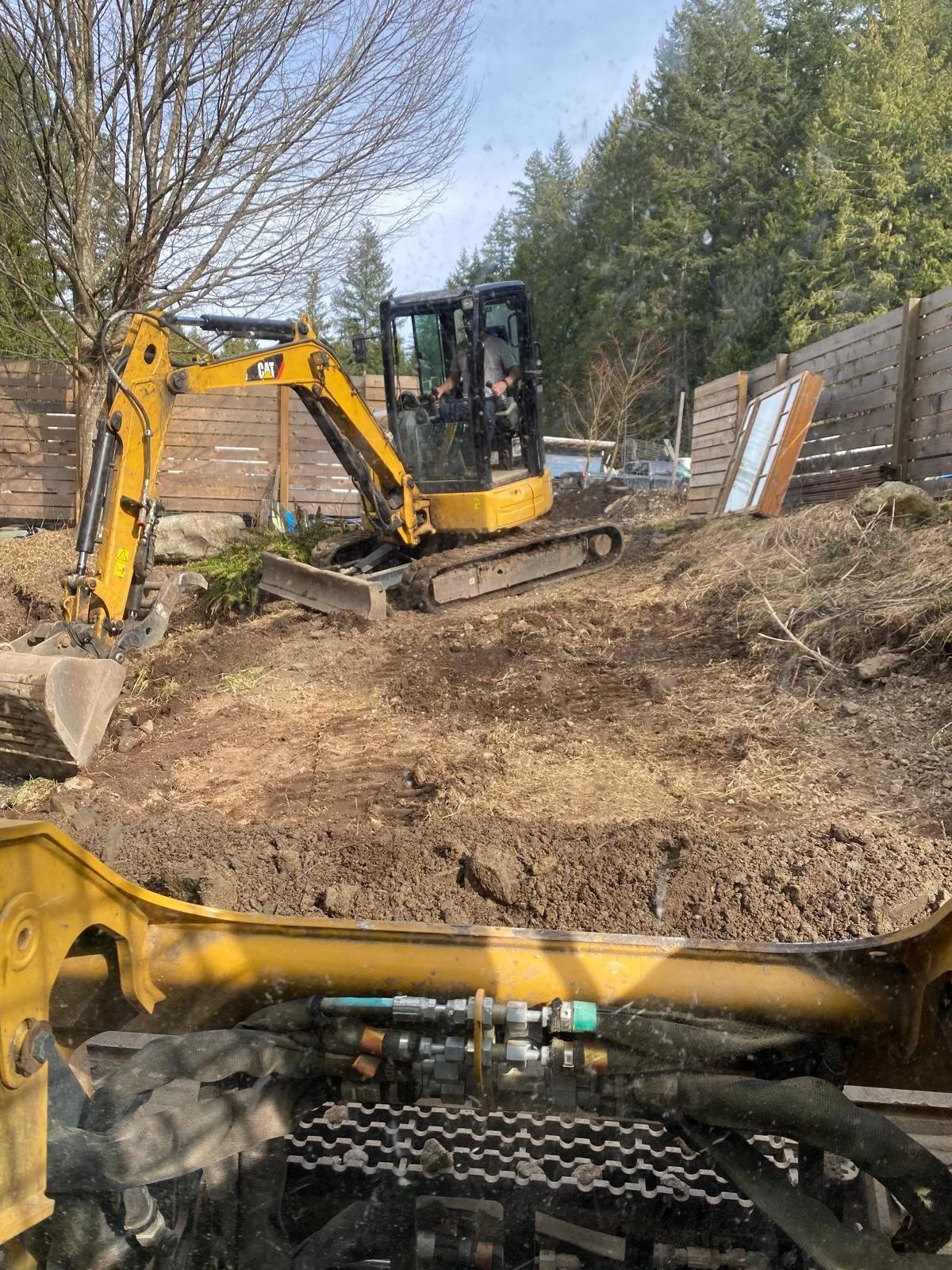 A construction site with a yellow Caterpillar excavator digging into dirt, surrounded by wooden fencing and trees in the background.