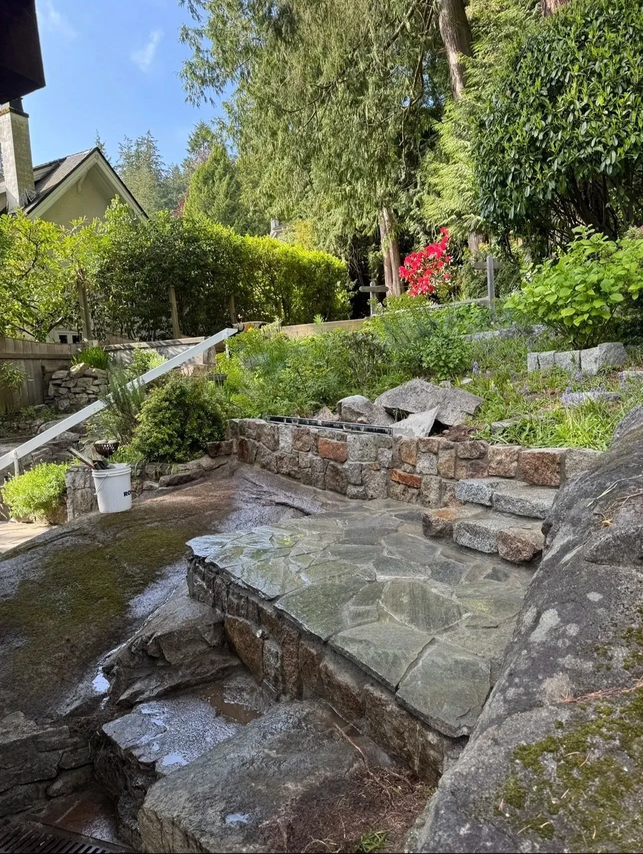 Backyard with a small waterfall flowing over large stones, surrounded by lush green plants and bushes, with a stone retaining wall and trees in the background.