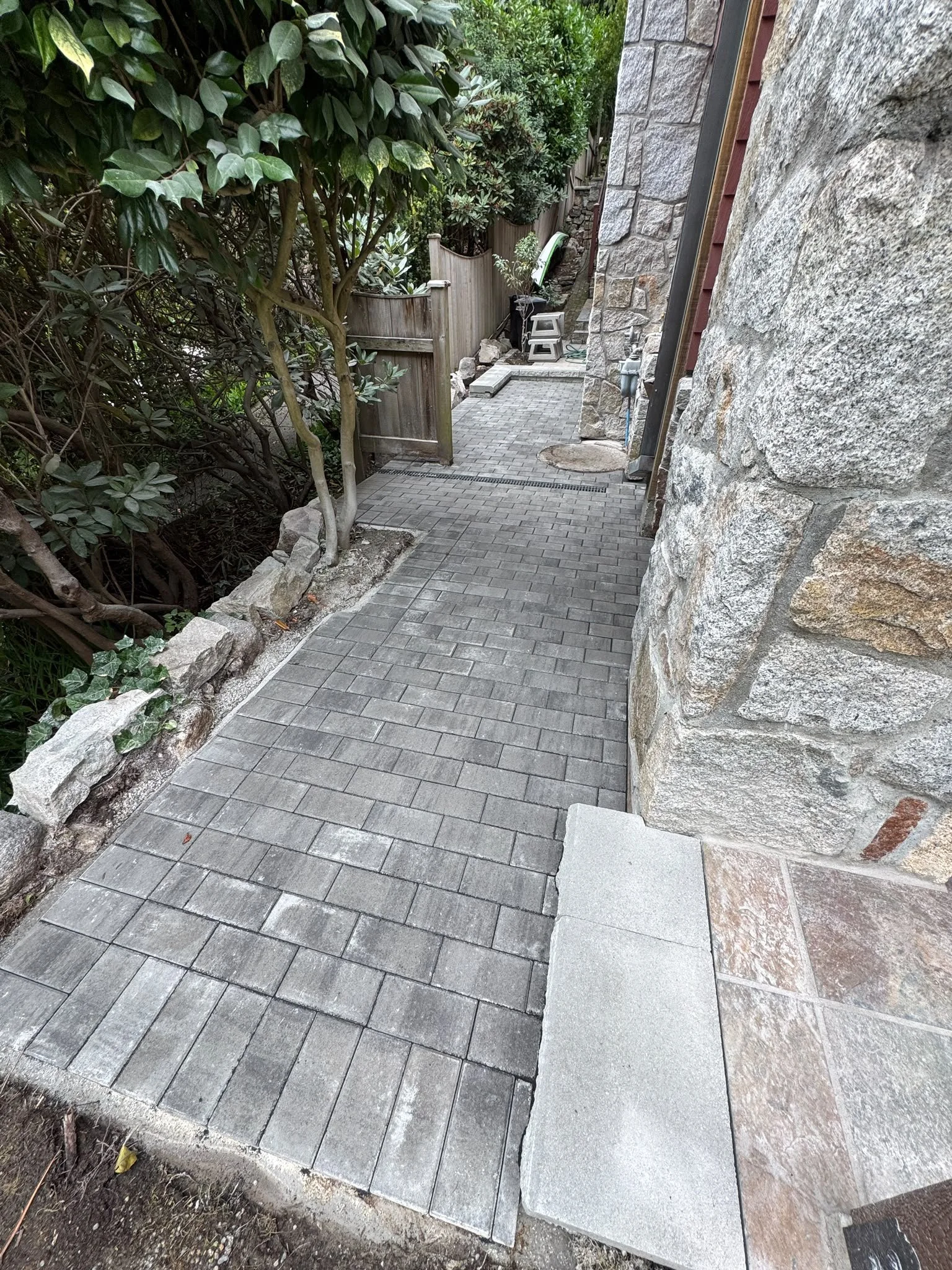 A narrow outdoor pathway with gray brick pavers, bordered by lush green bushes on the left and a stone building wall on the right. A small wooden fence and some gardening tools are visible in the background.