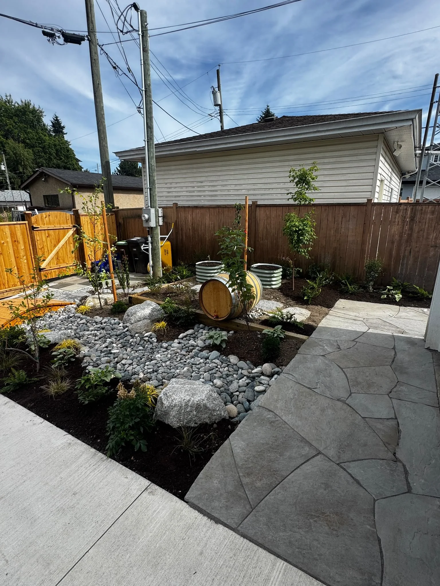 A backyard garden with a wooden fence, small trees, and landscaped rocks next to a concrete patio and a stone walkway. There are utility poles and wires in the background under a partly cloudy sky.