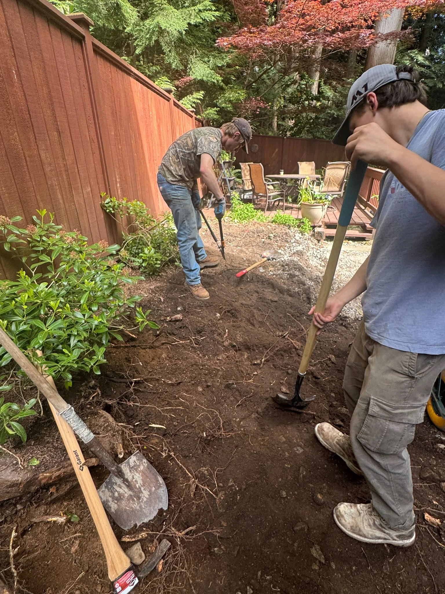 Two men working in a backyard garden, digging soil with shovels. One wears a cap and camouflage t-shirt, the other wears a gray baseball cap and gray cargo pants. There is a red wooden fence, trees with green and red leaves, and outdoor patio furnitu
