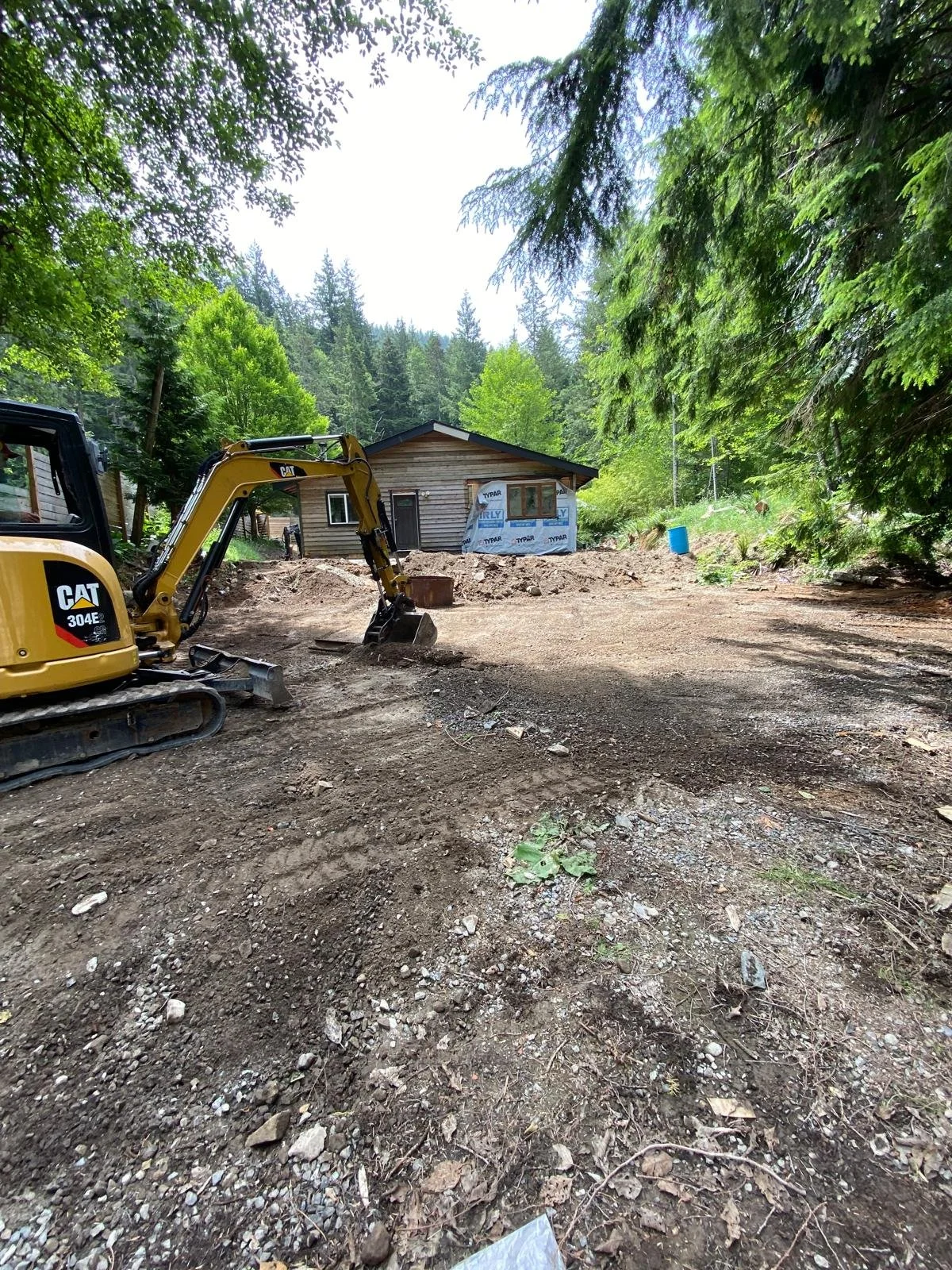 A construction site in a forested area with a small wooden house in the background, a yellow bulldozer parked on the dirt, and trees surrounding the site.