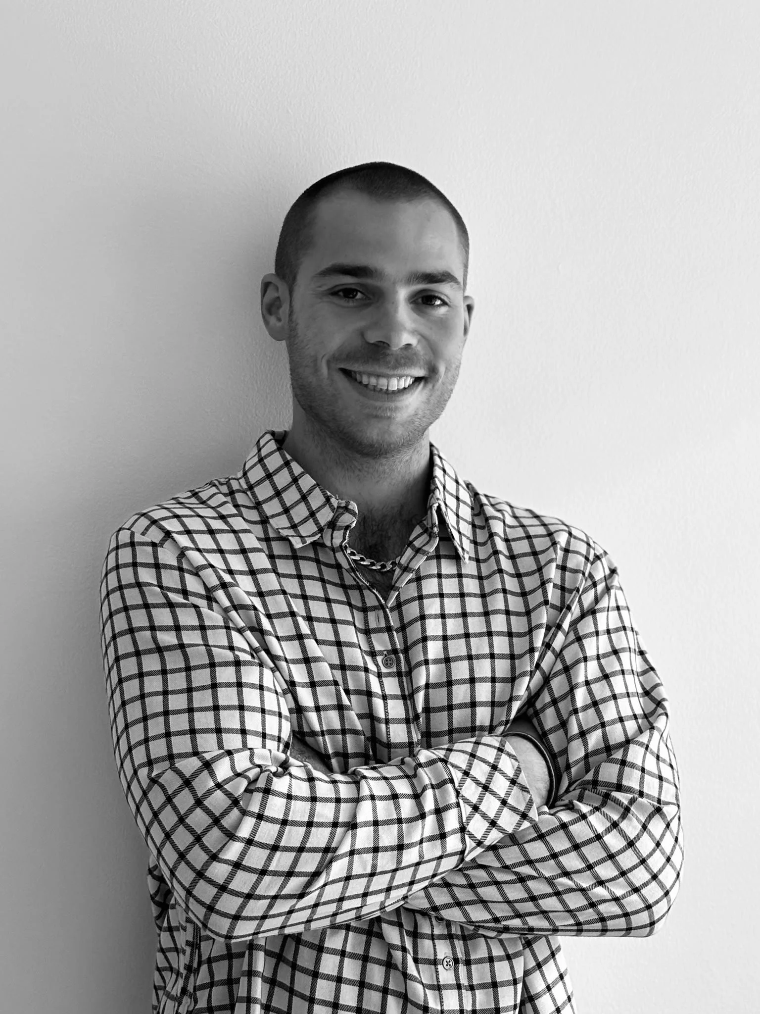 Black and white photo of a smiling man with short hair, wearing a checkered shirt with rolled-up sleeves and a chain necklace, standing with arms crossed against a plain wall.