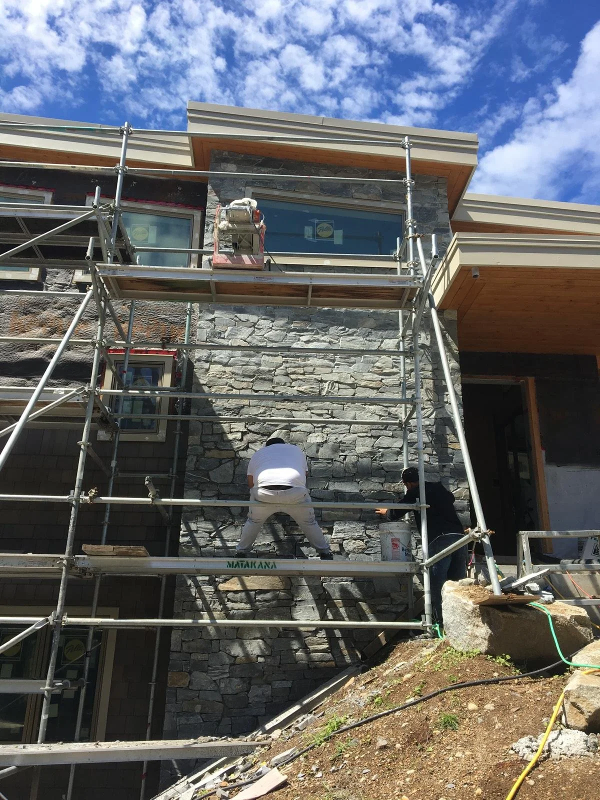 Construction workers installing stone veneer on the exterior of a building during daytime, with scaffolding and tools.