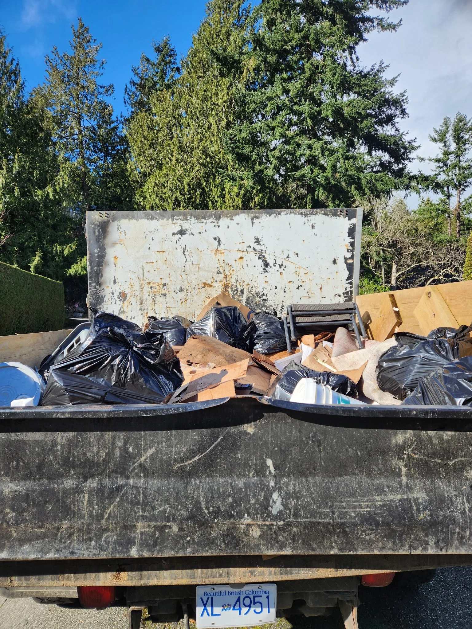 The back of a pickup truck filled with black trash bags, cardboard, and various debris, with a curved wooden fence and lush green trees in the background, and a British Columbia license plate visible on the truck.