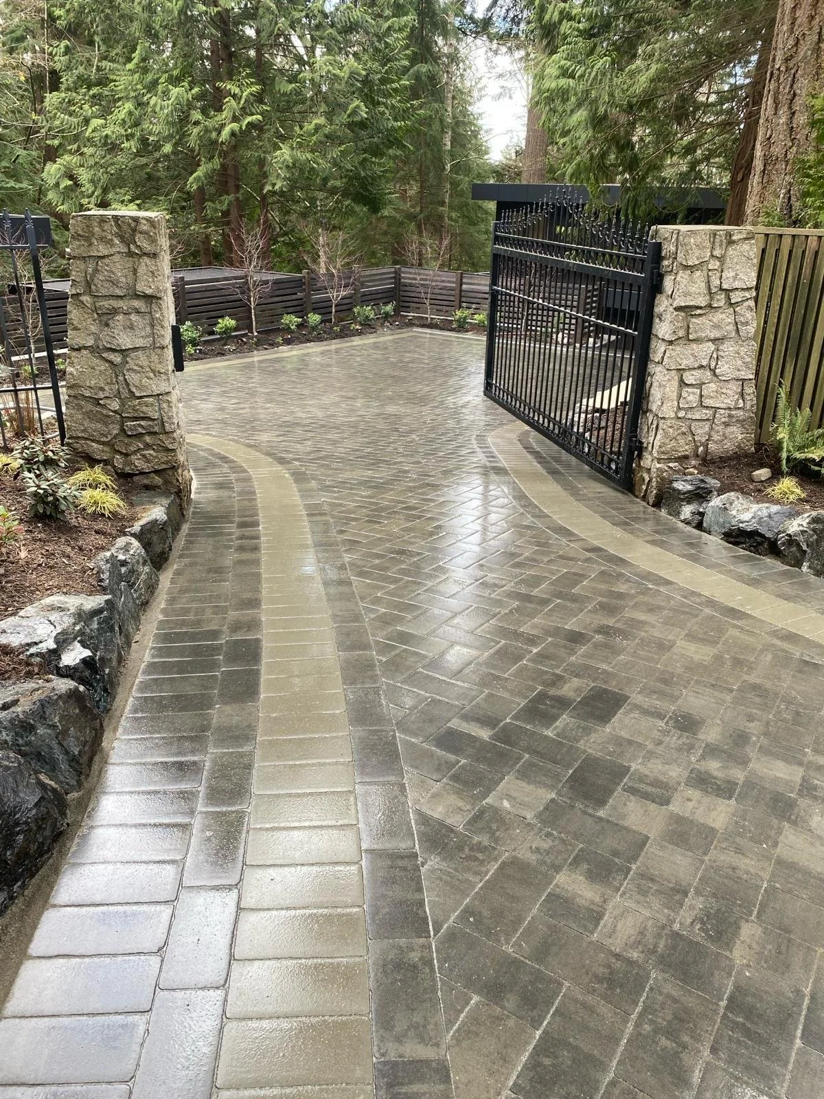 Photo of a paved driveway with a decorative border, surrounded by trees and plants, with a black iron gate supported by stone pillars.