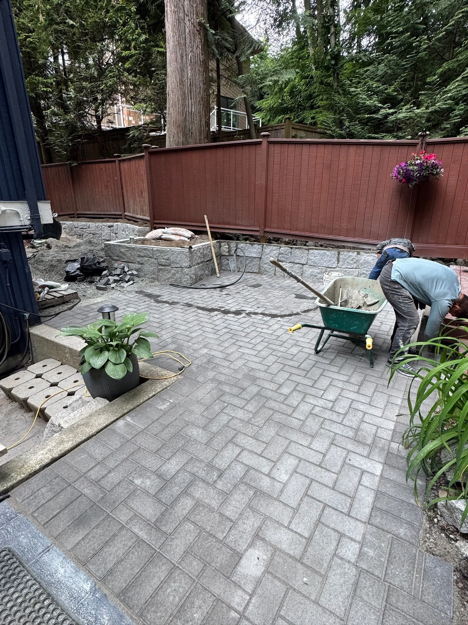 Two workers installing paver bricks in a backyard patio area near a wooden and stone retaining wall. The area is surrounded by tall trees and a fence, with a hanging flower basket visible on the right.