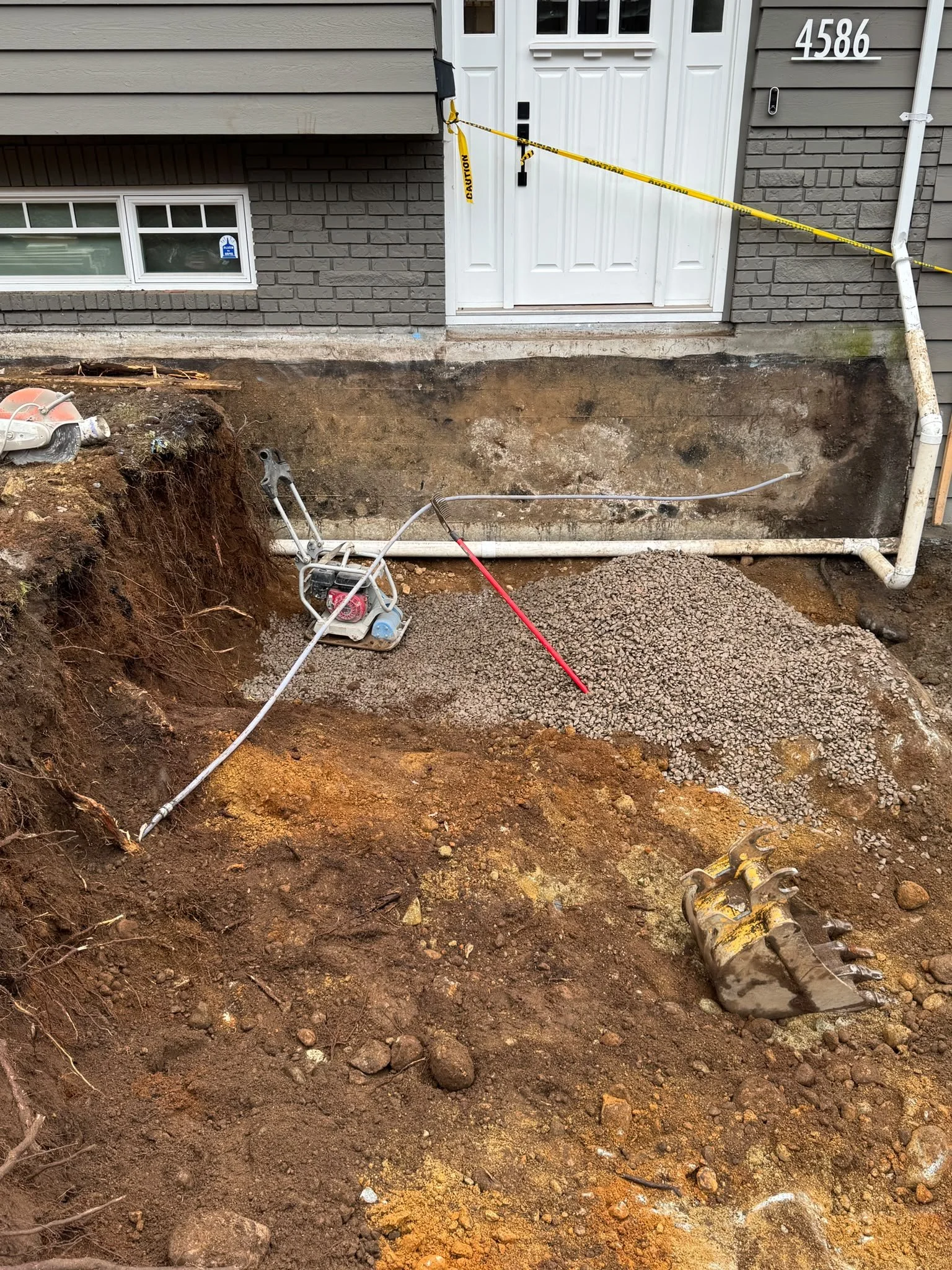 Underground plumbing installation and excavation site in front of a house, visible pipes, dirt, and construction equipment.