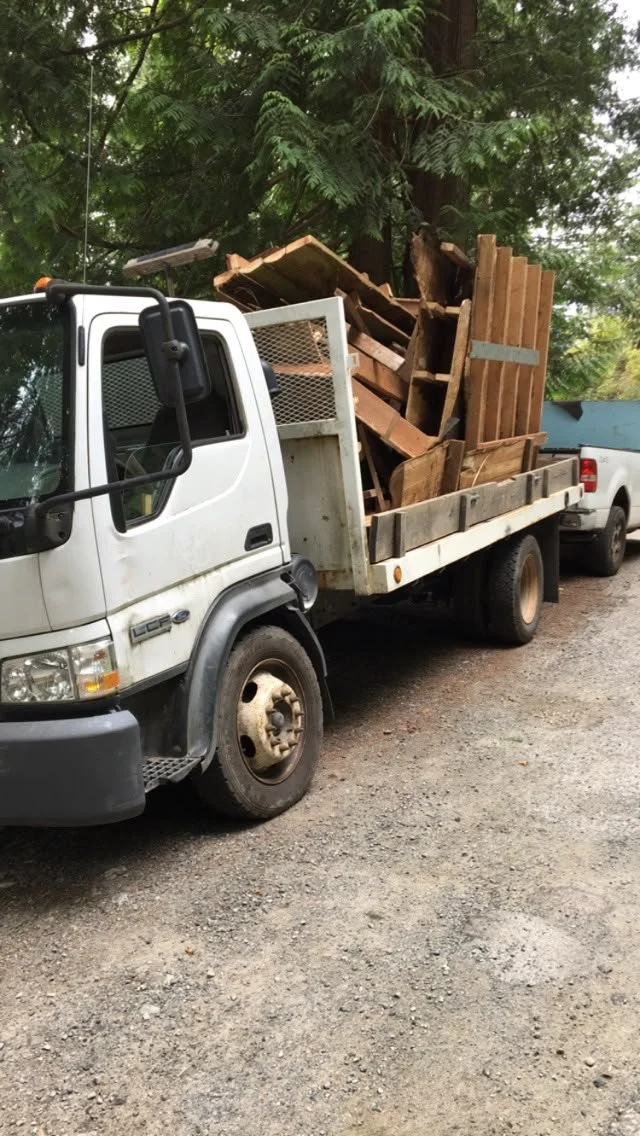 A white flatbed truck loaded with wooden debris, parked on a gravel lot with trees in the background.