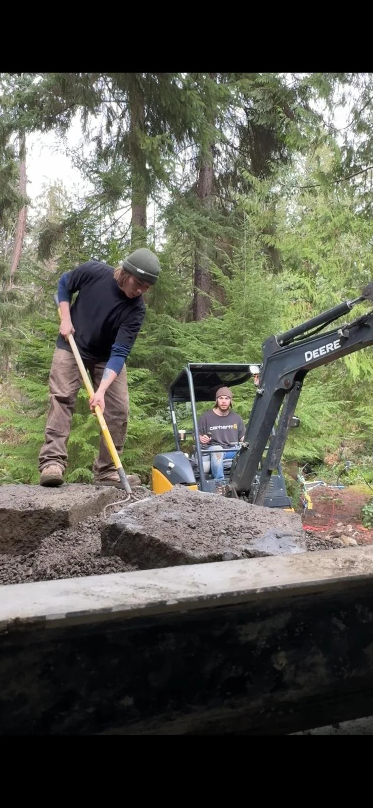 Two men working outdoors in a forest, one using a rake on a dirt surface and the other sitting in a small backhoe.